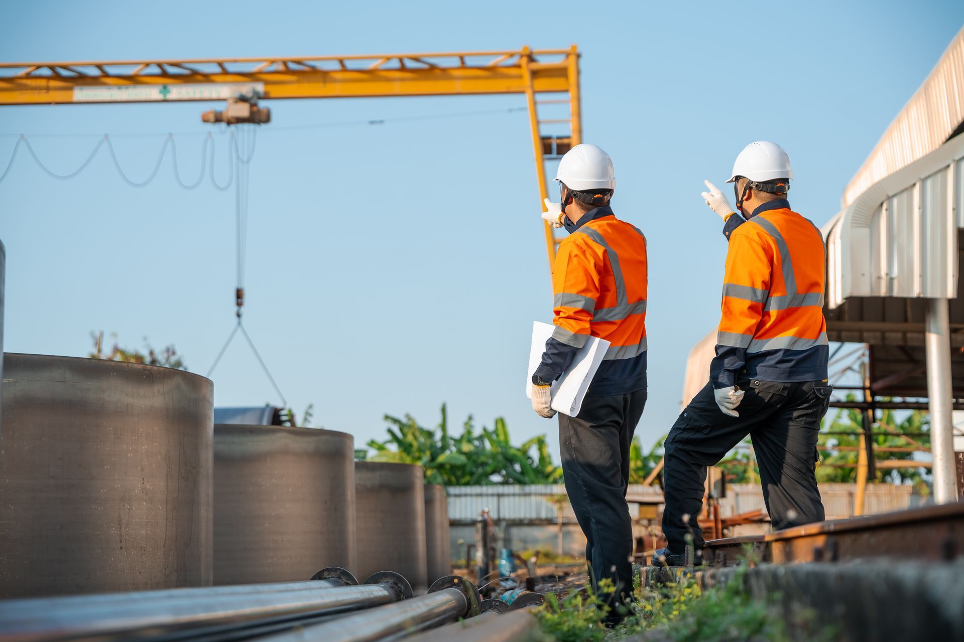 Two construction workers in orange vests and white hard hats stand beside a crane at a worksite.
