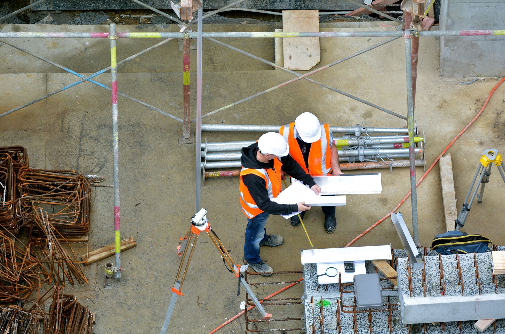 Two construction workers in orange safety vests and hard hats review plans at a worksite.