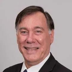 Headshot of a person with short brown hair, wearing a dark suit jacket and a white collared shirt against a gray background.
