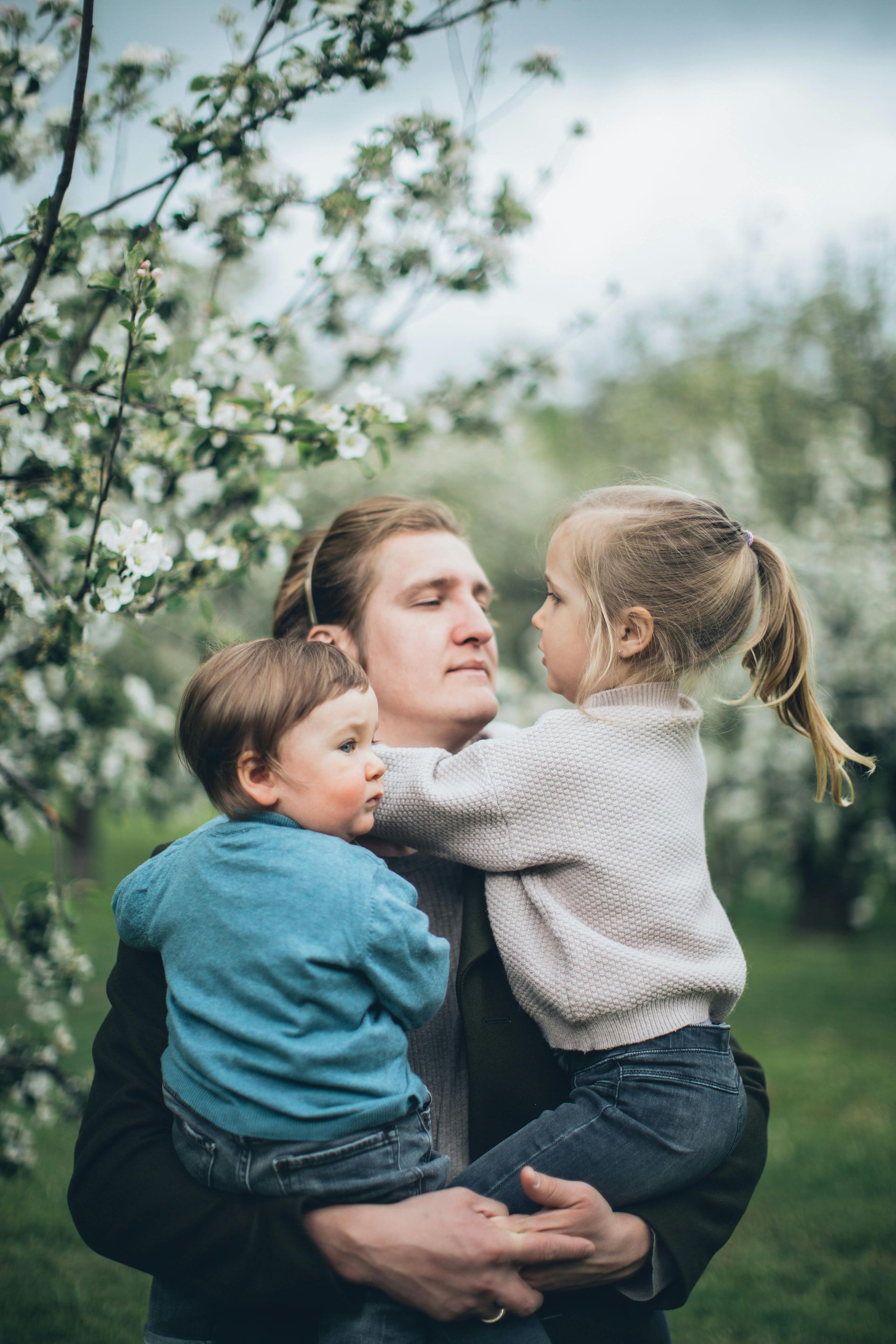 A person holds two children in their arms in an orchard filled with white blooming trees.