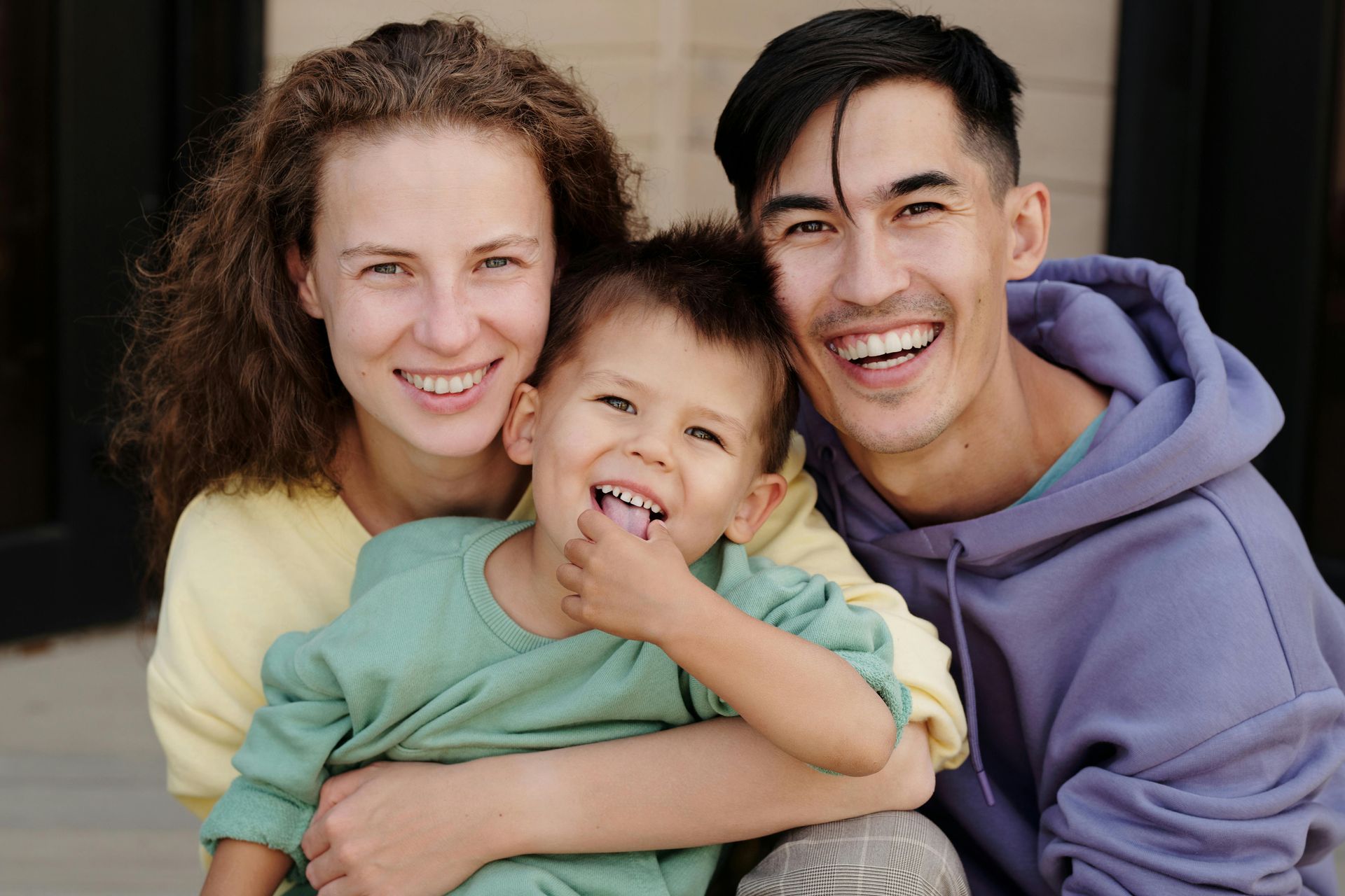 A happy family of three—a woman, a man, and a young child—smiling closely together outdoors.