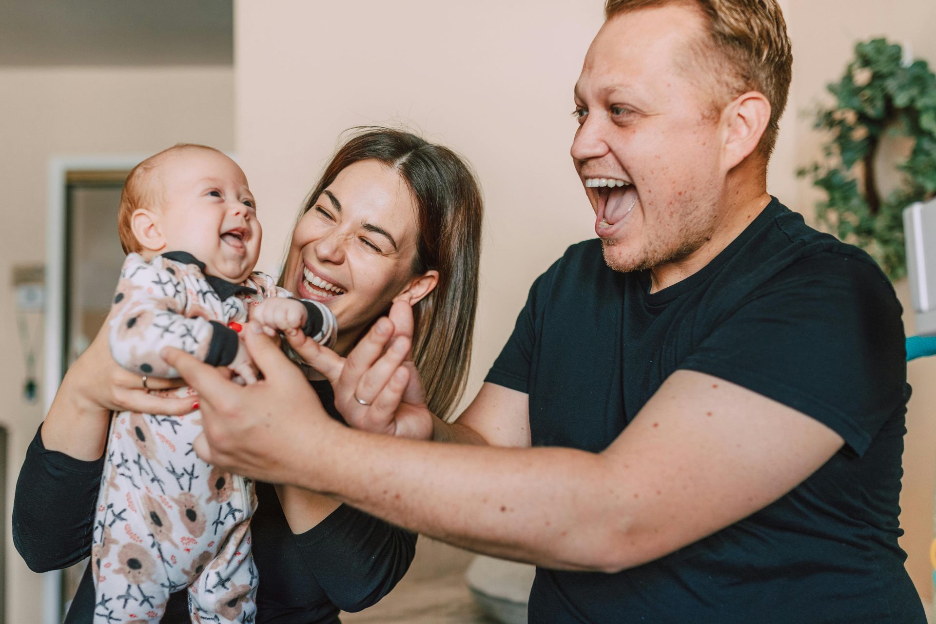 A smiling person holds a laughing infant while another person leans in toward the baby with an excited, joyful expression.