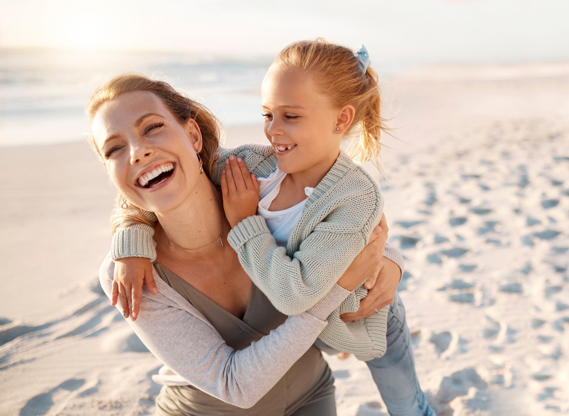 A smiling woman gives a child a piggyback ride on a sandy beach during the day.