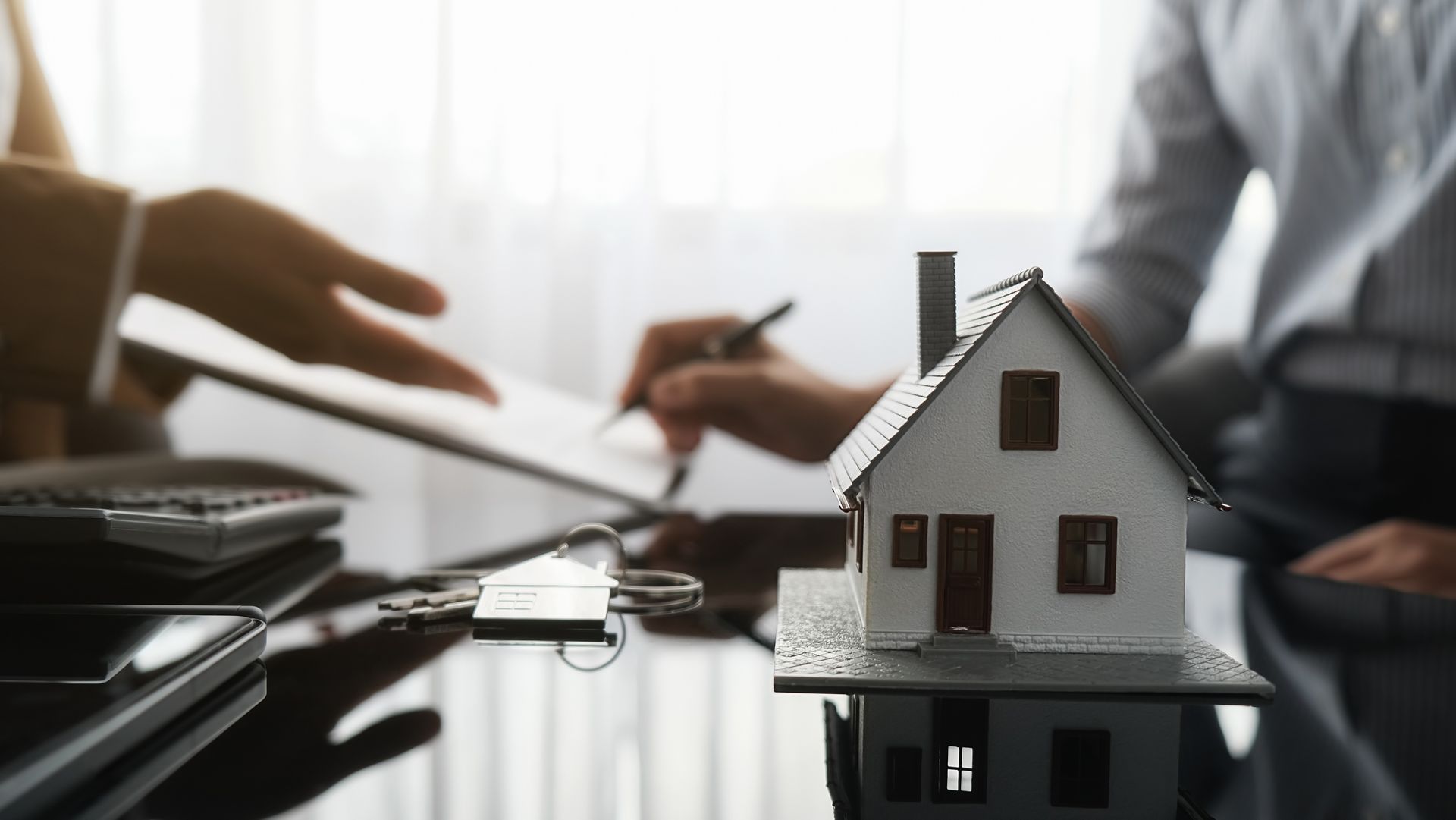 Small model house on a table with people discussing and signing papers in the background