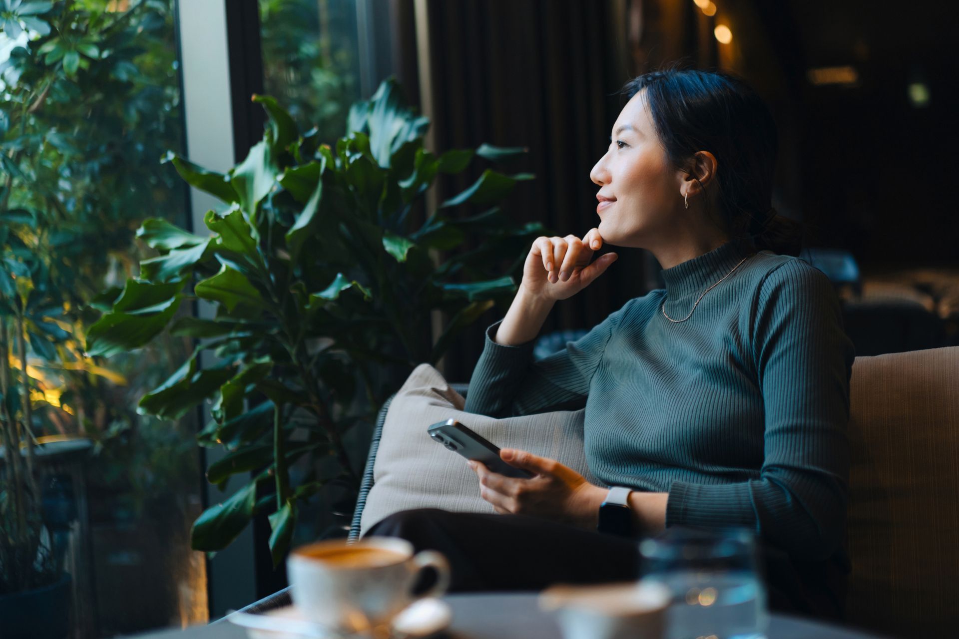 Woman gazing out a window, holding a phone, with coffee and plants nearby.