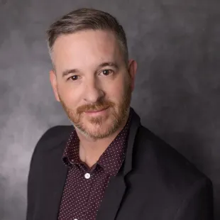 Man with salt-and-pepper hair, wearing a dark blazer over a maroon dotted shirt, smiling at the camera.