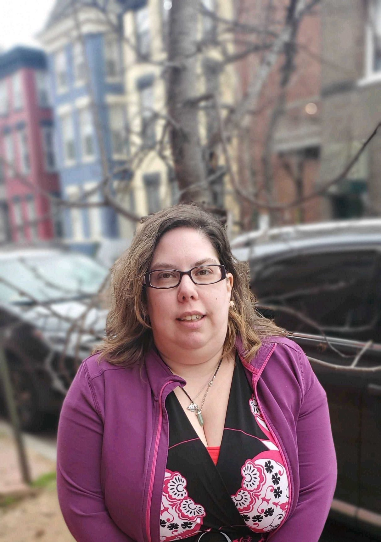 A woman wearing glasses and a purple jacket is standing in front of a building.