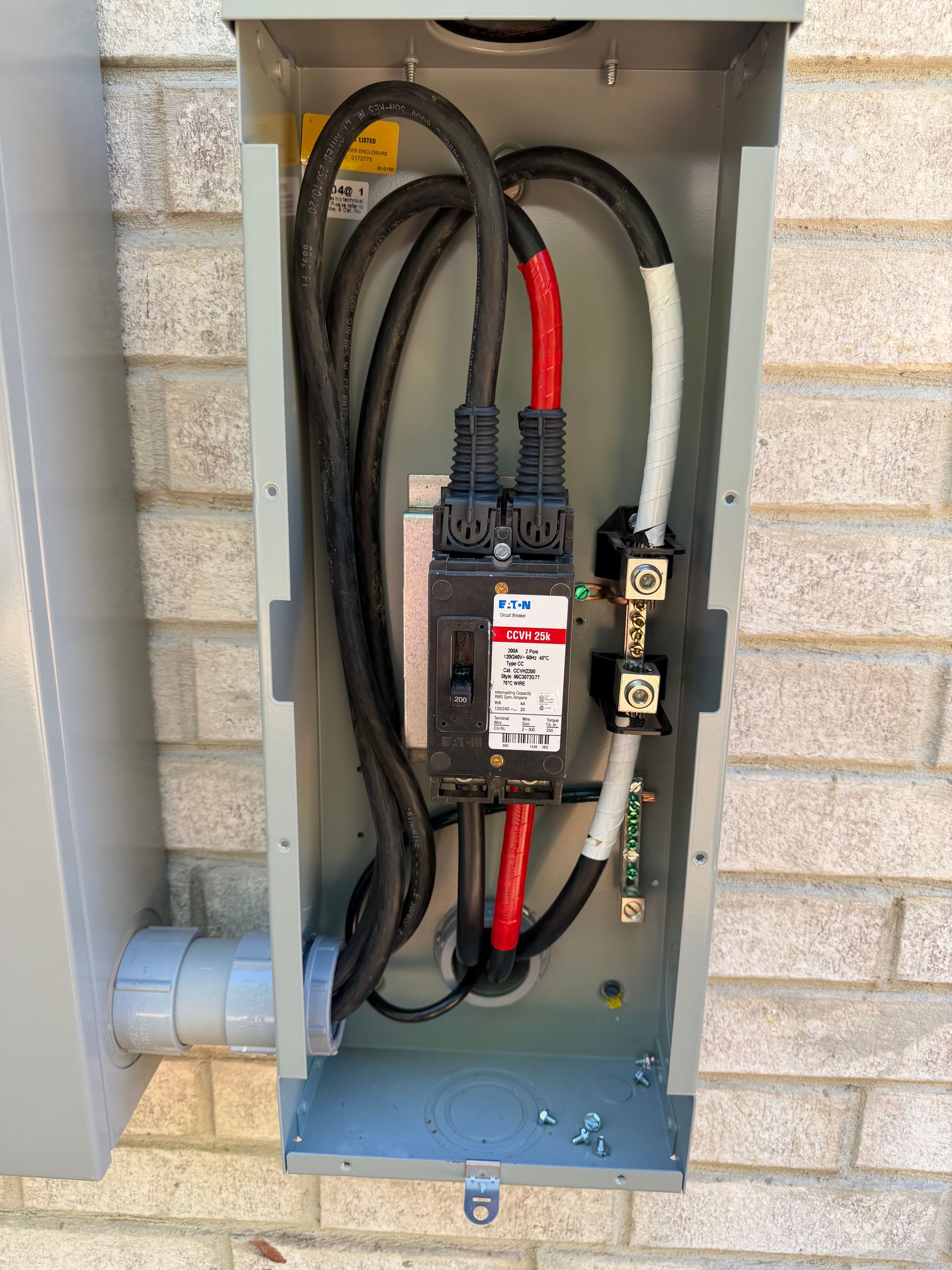 Open electrical breaker panel with thick black and red cables inside a gray metal box on a brick wall
