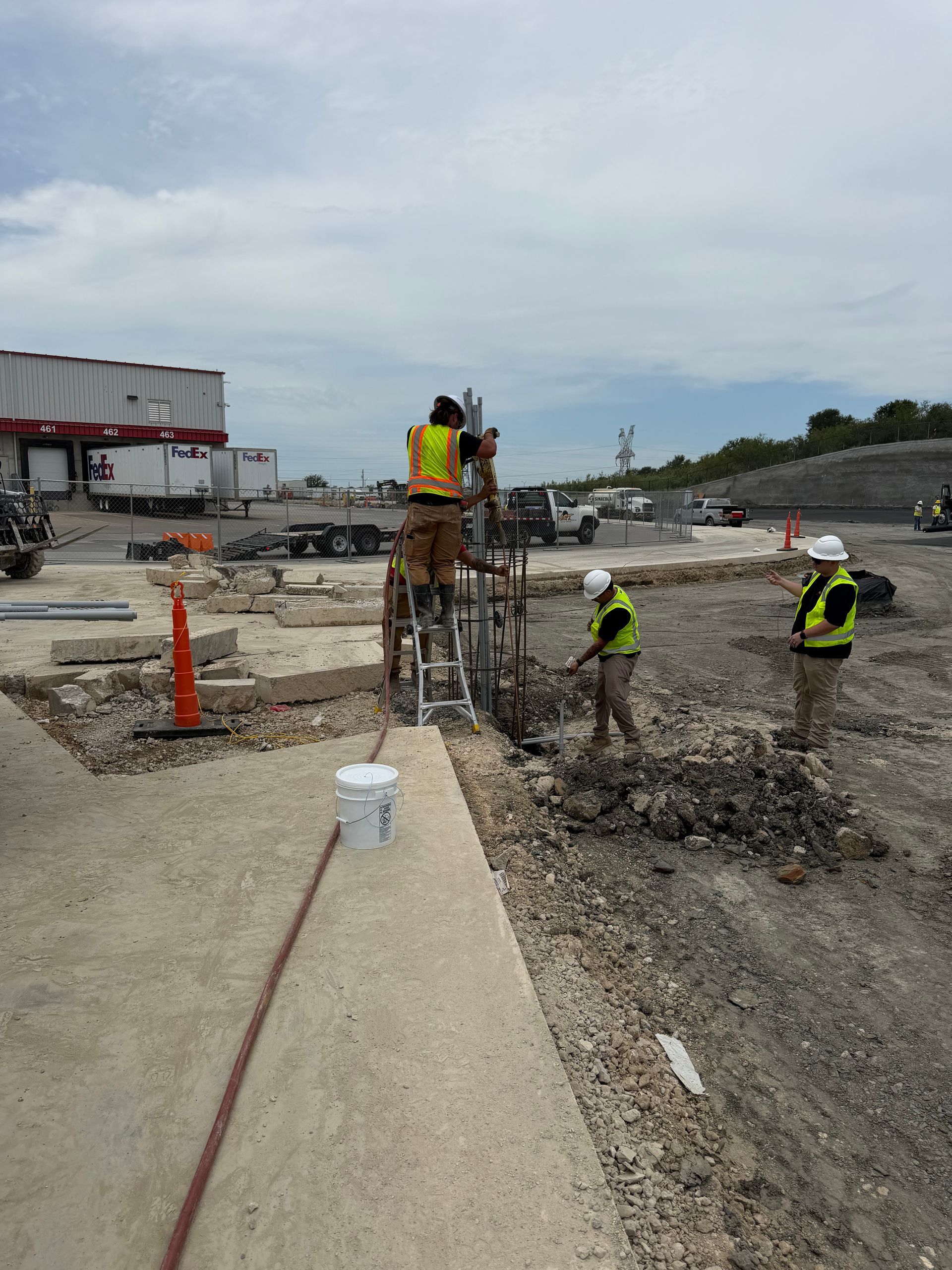Construction workers pouring concrete in a road trench beside orange cones and heavy machinery.