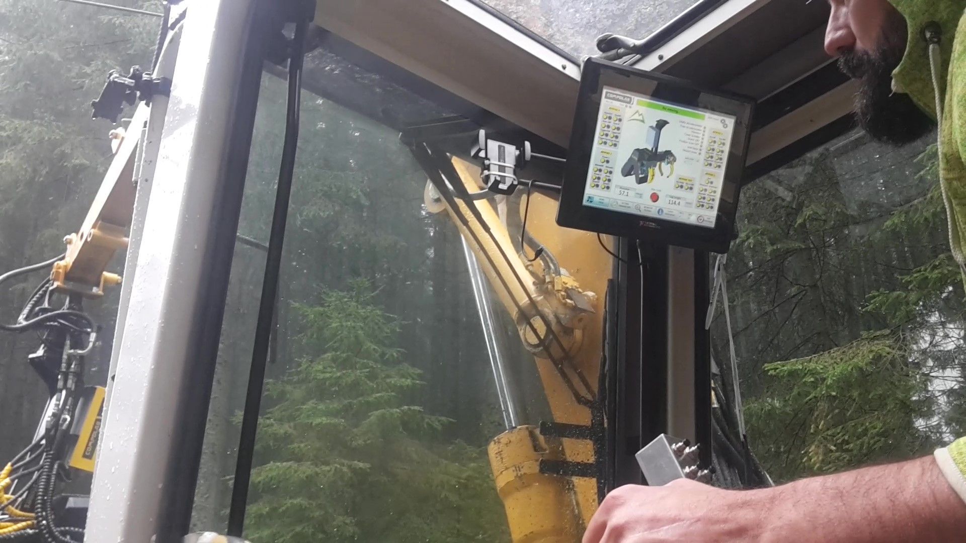 All'interno della cabina dell'escavatore: vista dell'operatore sui comandi e sullo schermo che visualizza le funzioni della macchina, foresta visibile all'esterno.