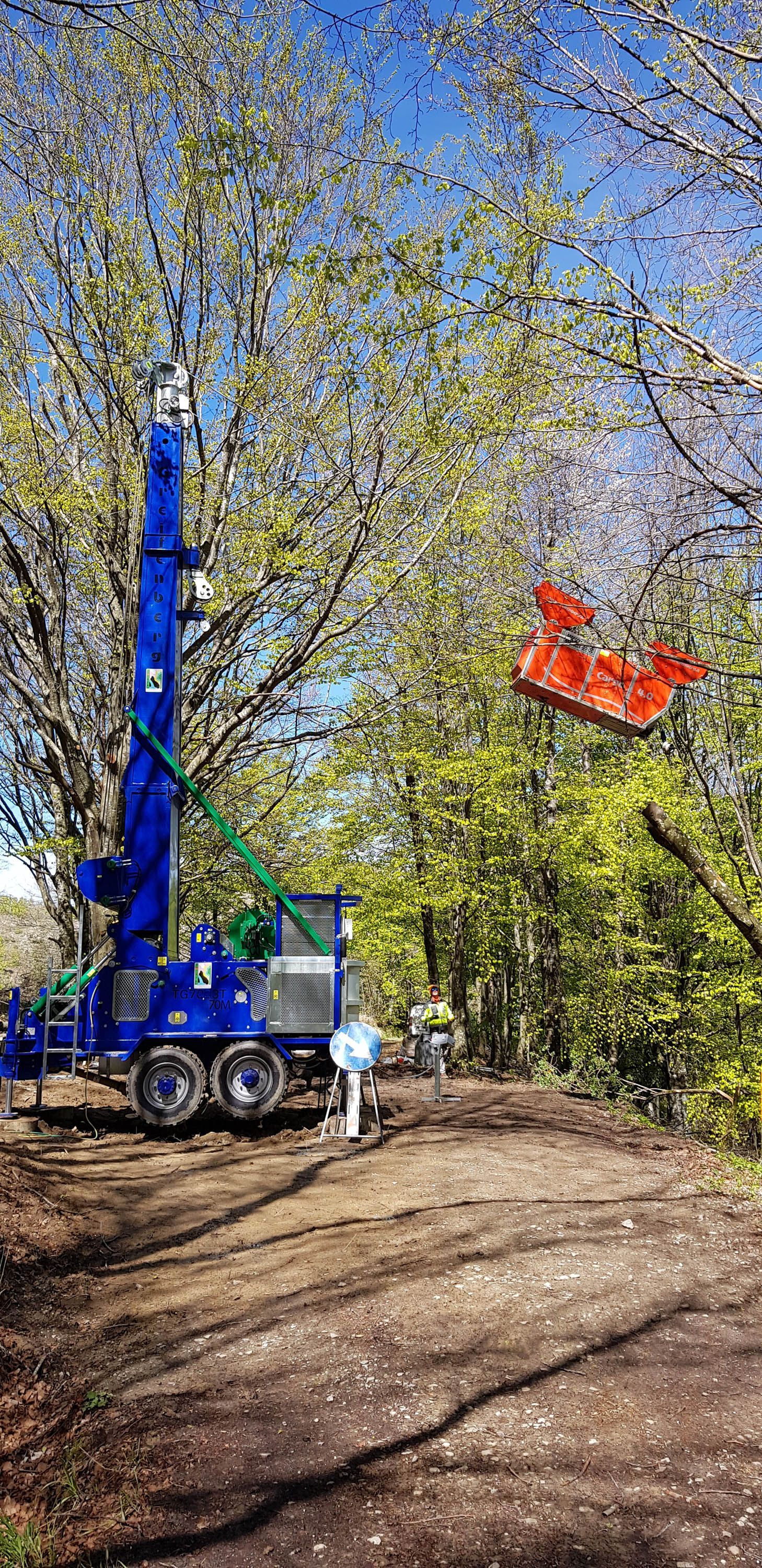 Una piattaforma di perforazione blu in una zona boscosa con una benna di sollevamento rossa tra gli alberi in una giornata di sole.