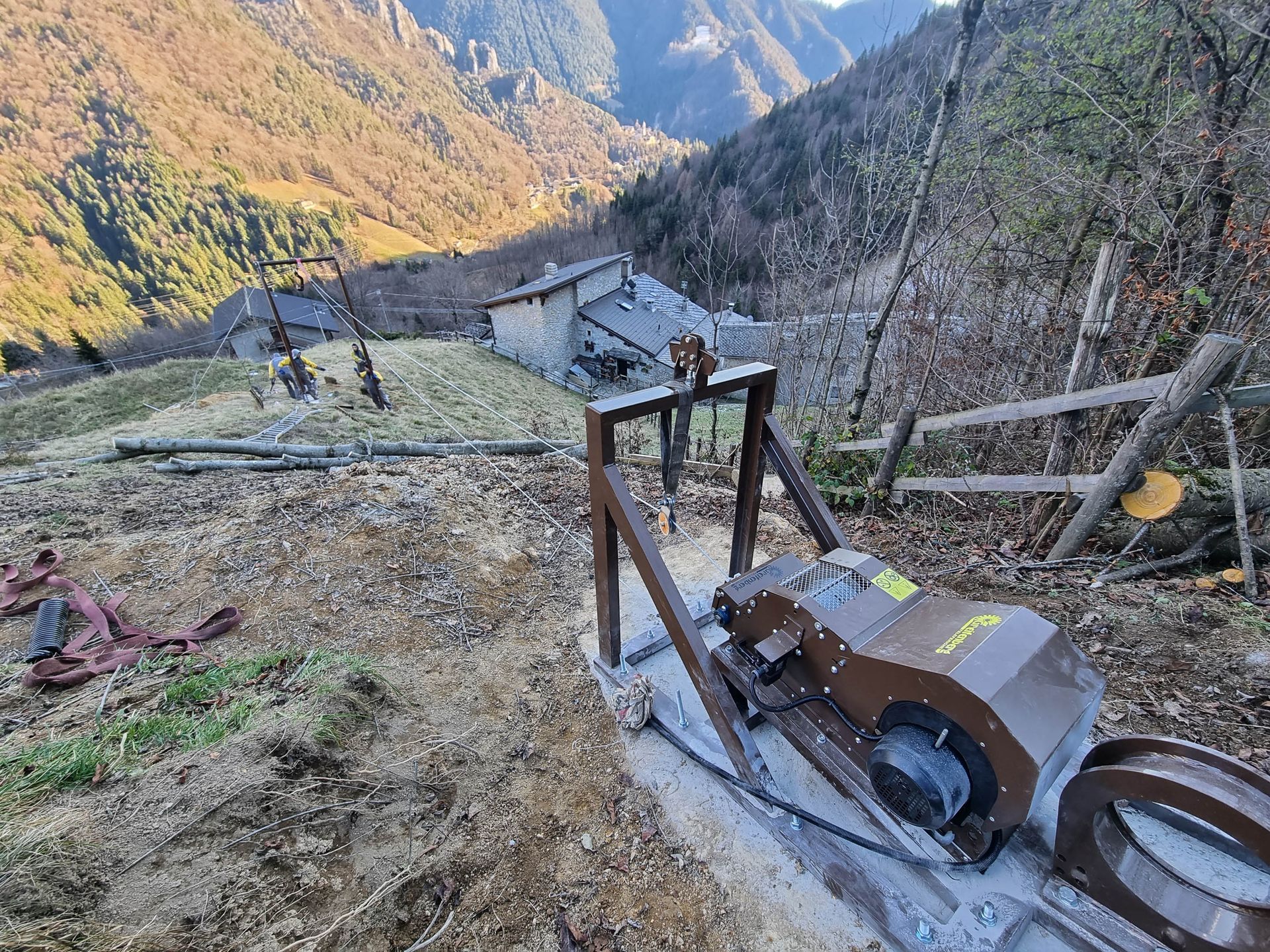Macchina che taglia il legno su una collina; sullo sfondo edifici in legno e montagne.