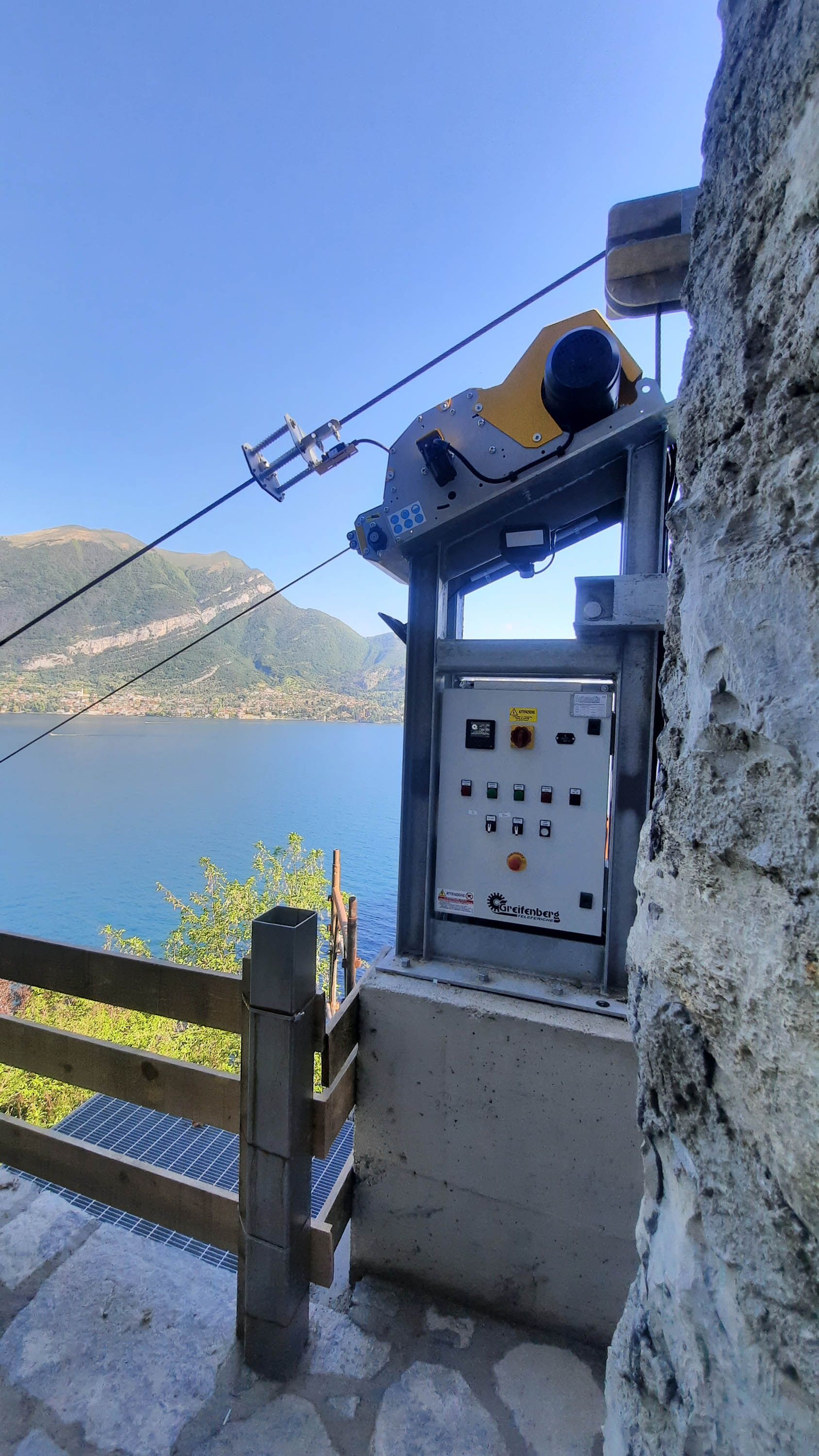 Stazione della funivia in riva a un lago, con pannello di controllo e macchinari. Cielo azzurro, muro di pietra e recinzione in legno.