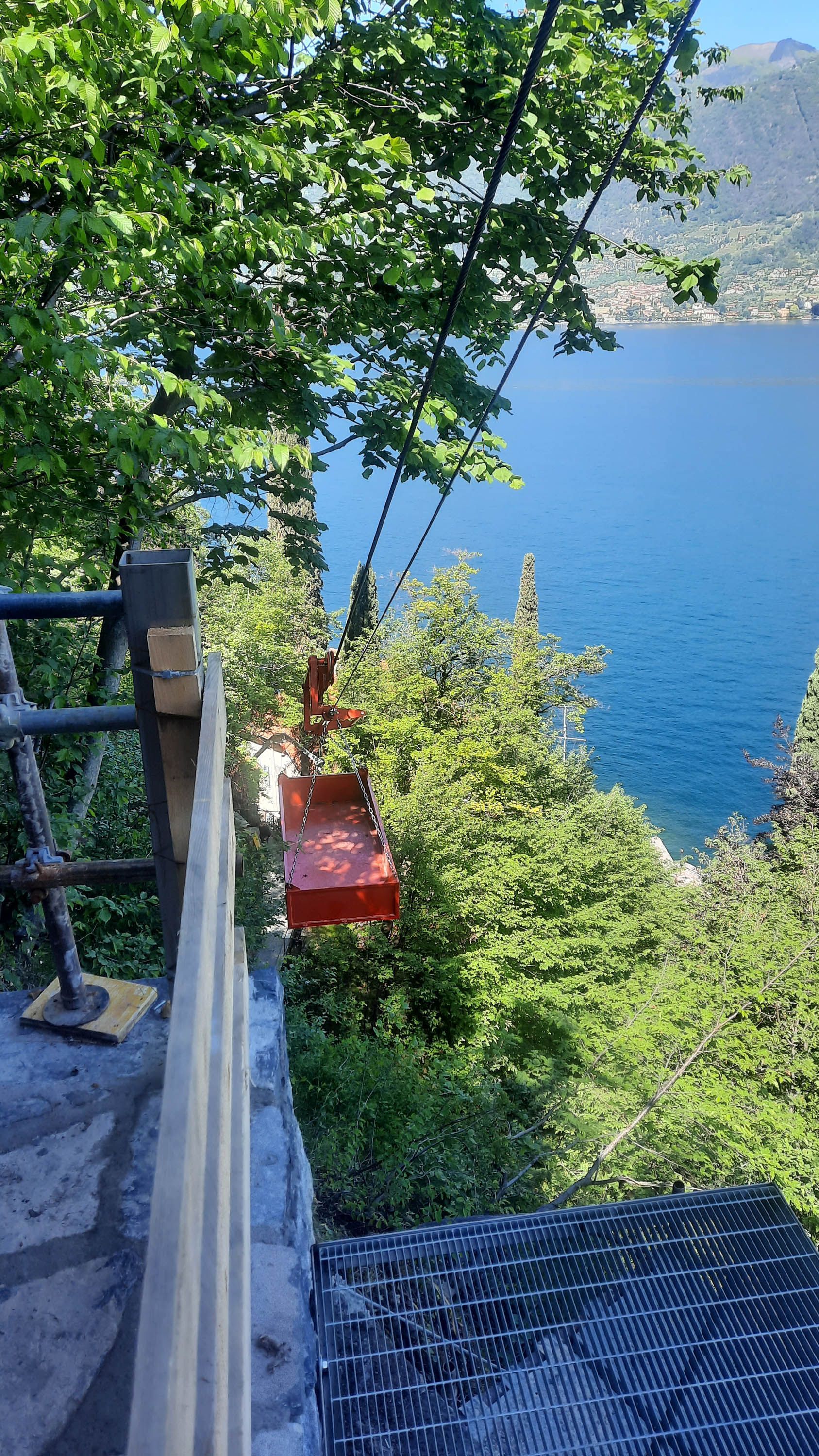Una seggiovia rossa attraversa un lago blu, circondato da alberi verdi e da un muro di pietra.