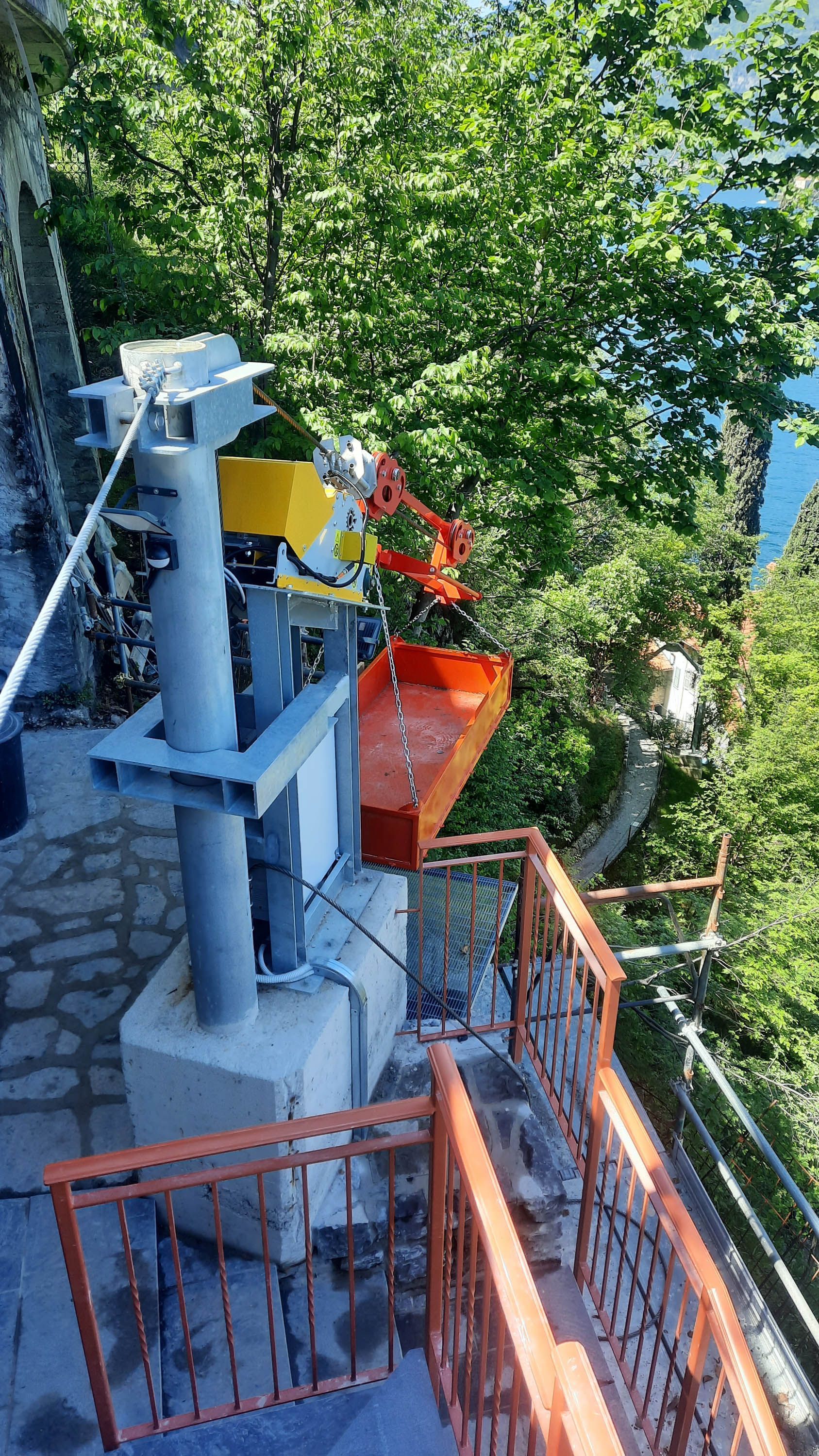 Una funivia con un secchio rosso e giallo, vicino a una ringhiera arancione, immersa in una foresta.
