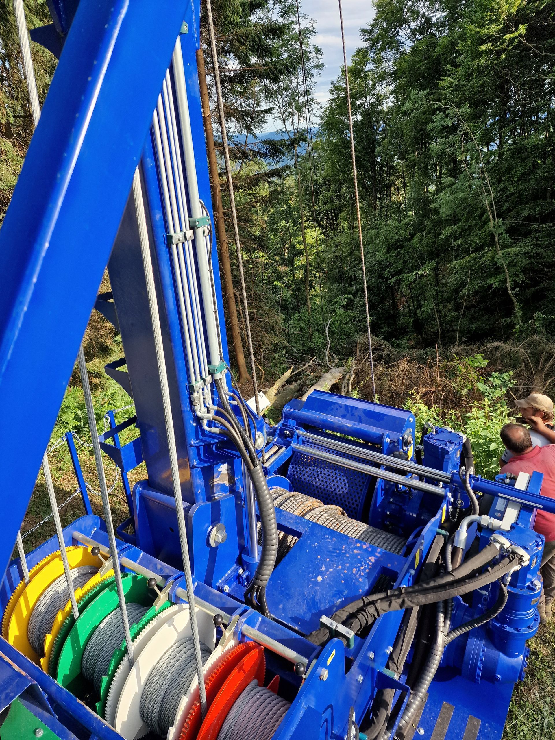 Piattaforma di perforazione blu in una foresta, con bobine di cavi colorati e due persone che osservano.