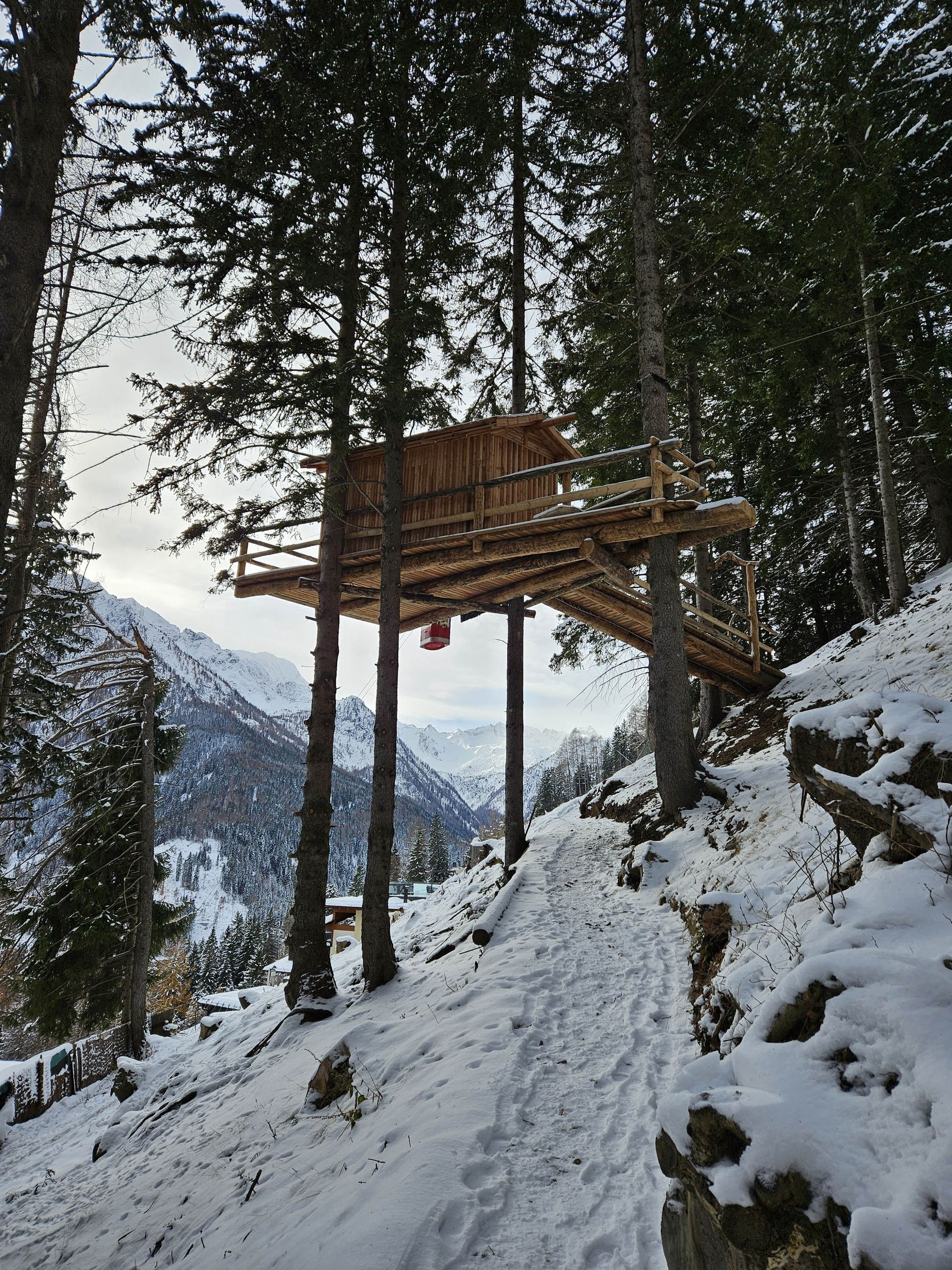Casa sull'albero in una foresta innevata, costruita su palafitte. Vi si accede tramite una passerella in legno. Montagne sullo sfondo.