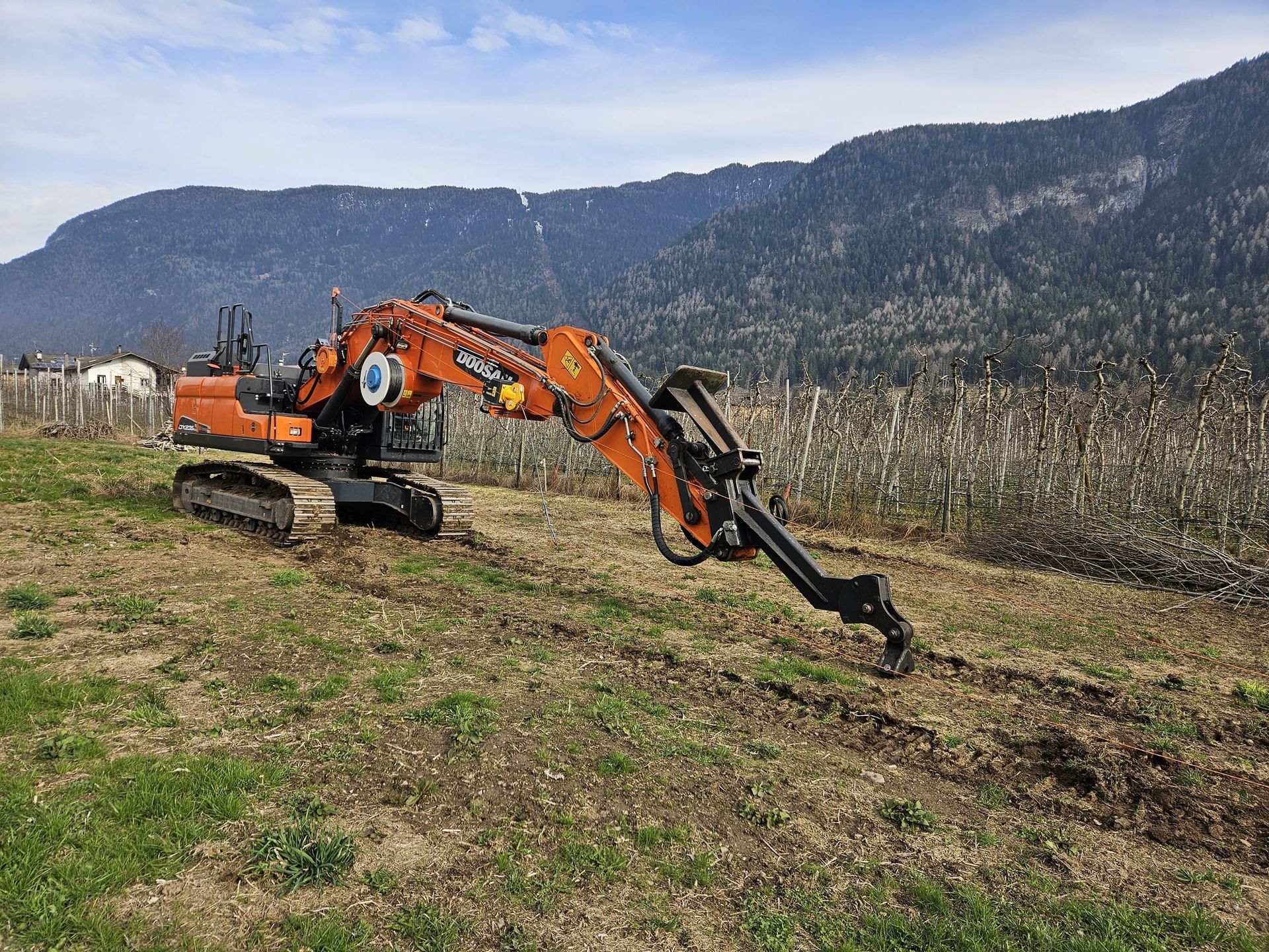 Escavatore arancione in un campo, al lavoro di fronte alle montagne, per ripulire la vegetazione.
