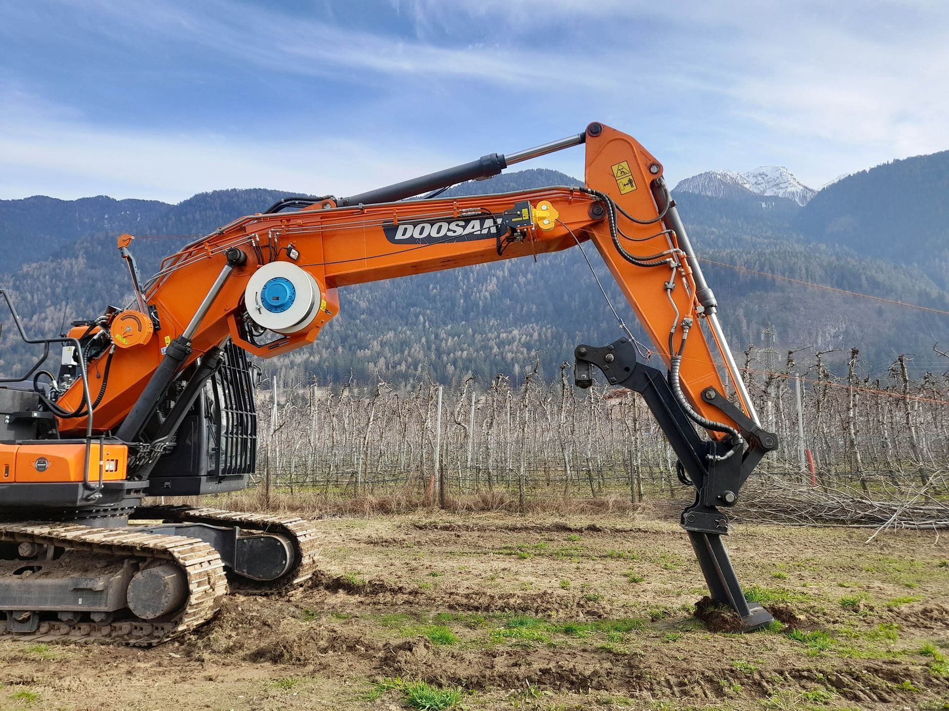 Escavatore Doosan arancione in un campo, con braccio esteso verso il basso. Montagne sullo sfondo.