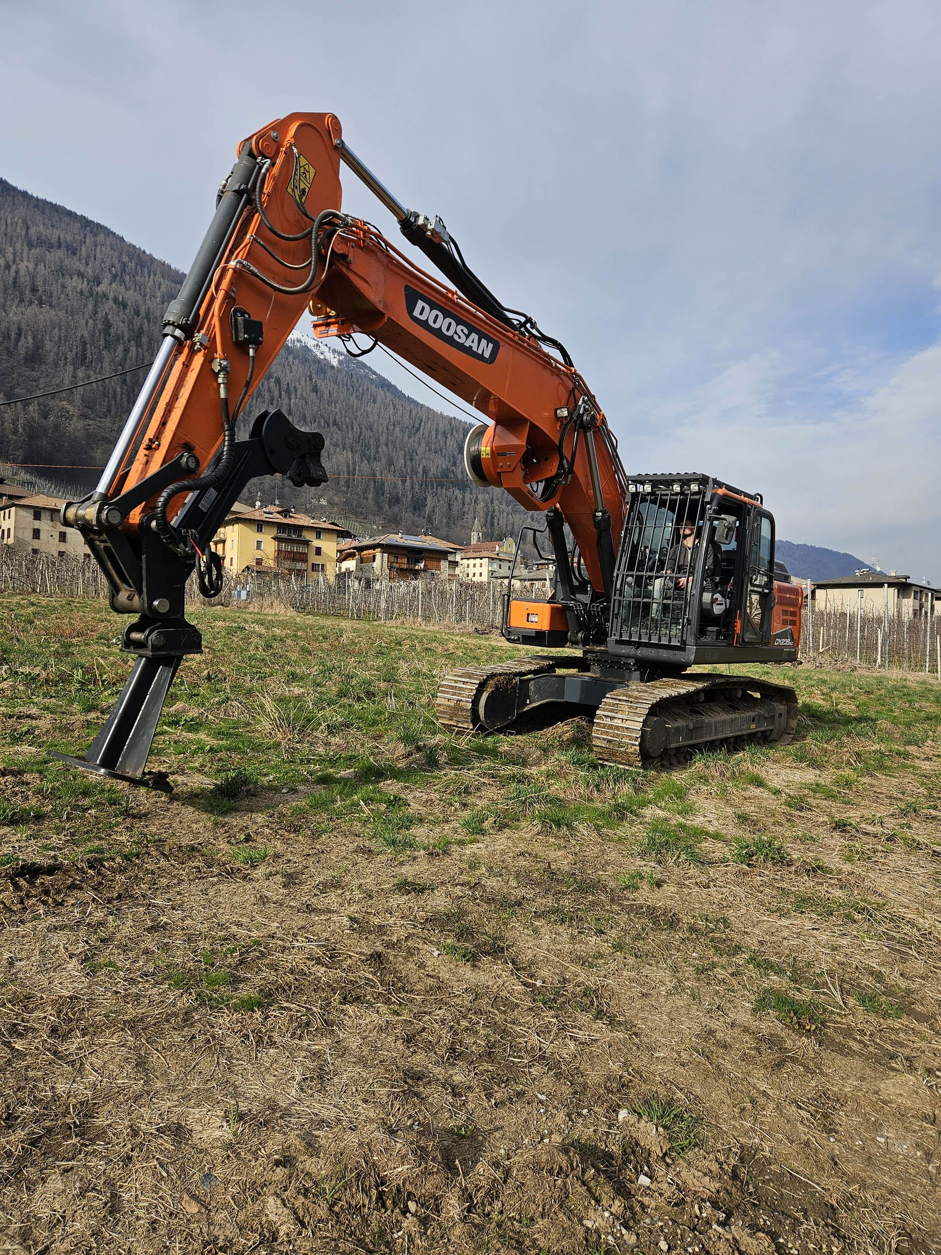 Escavatore Doosan arancione parcheggiato in un campo. Sullo sfondo alberi ed edifici sotto un cielo nuvoloso.