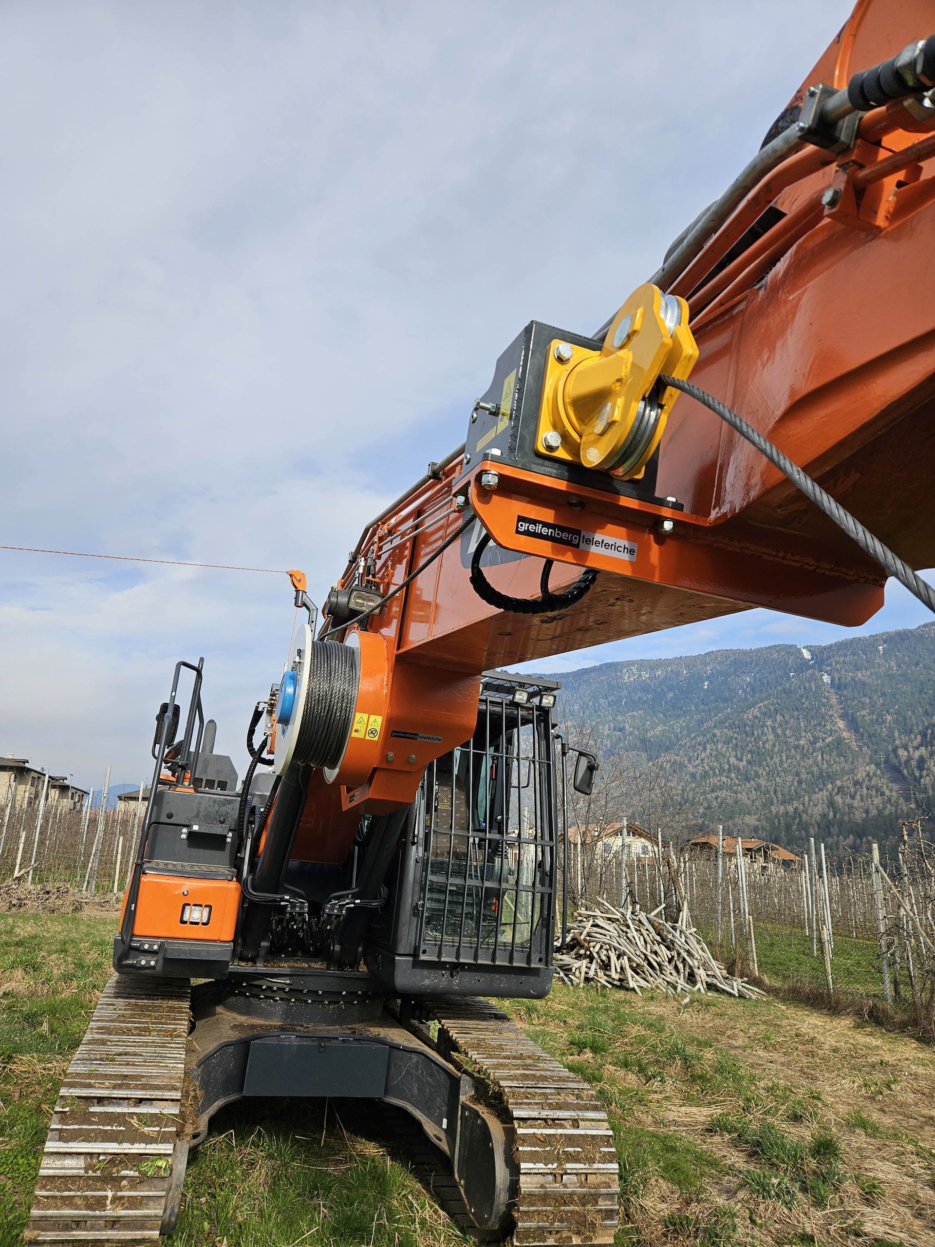 Escavatore arancione su cingoli, braccio esteso con verricello. Erba verde, cielo nuvoloso, montagne sullo sfondo.