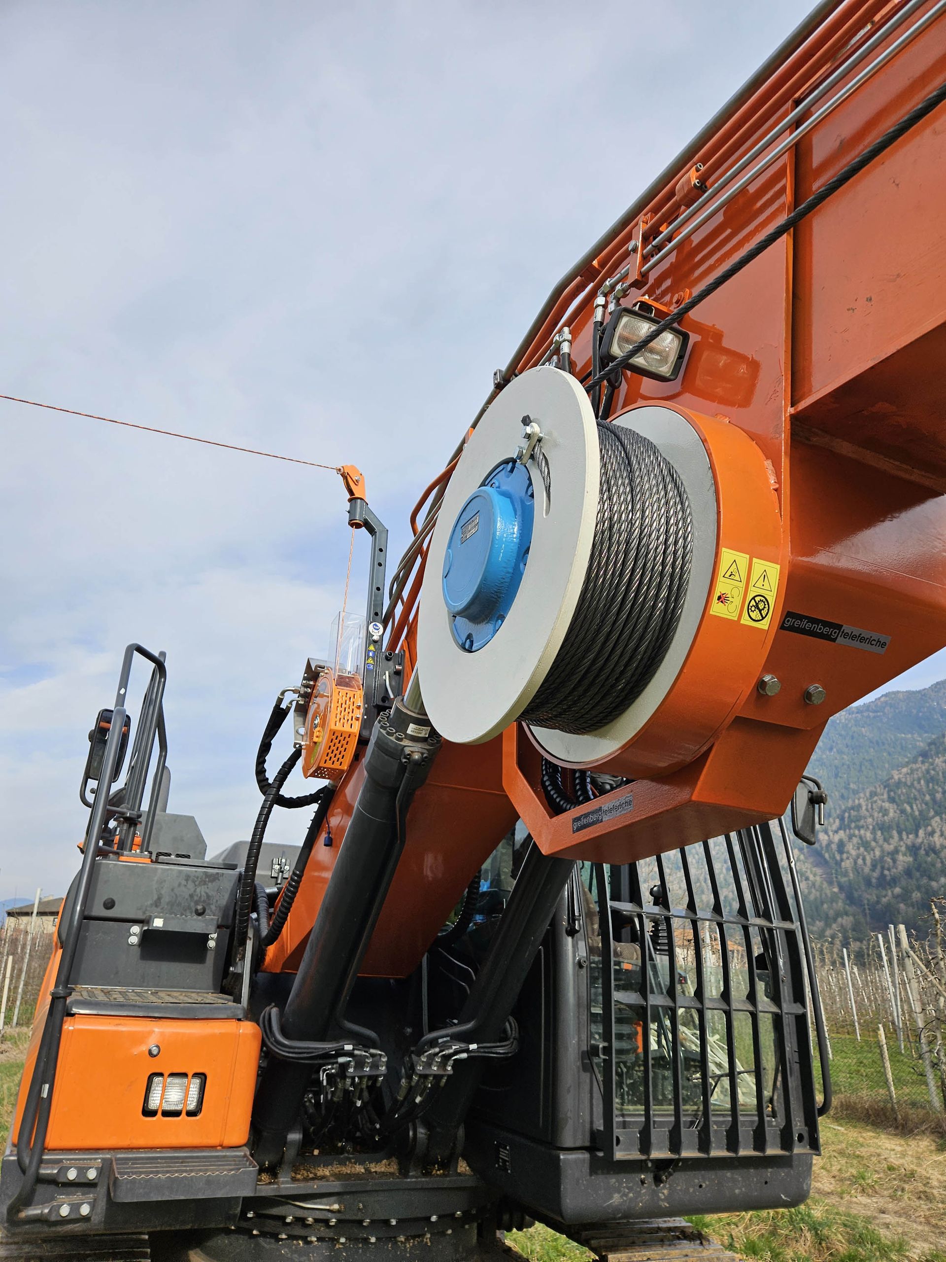 Escavatore arancione con verricello e cavo d'acciaio sullo sfondo di un cielo nuvoloso.