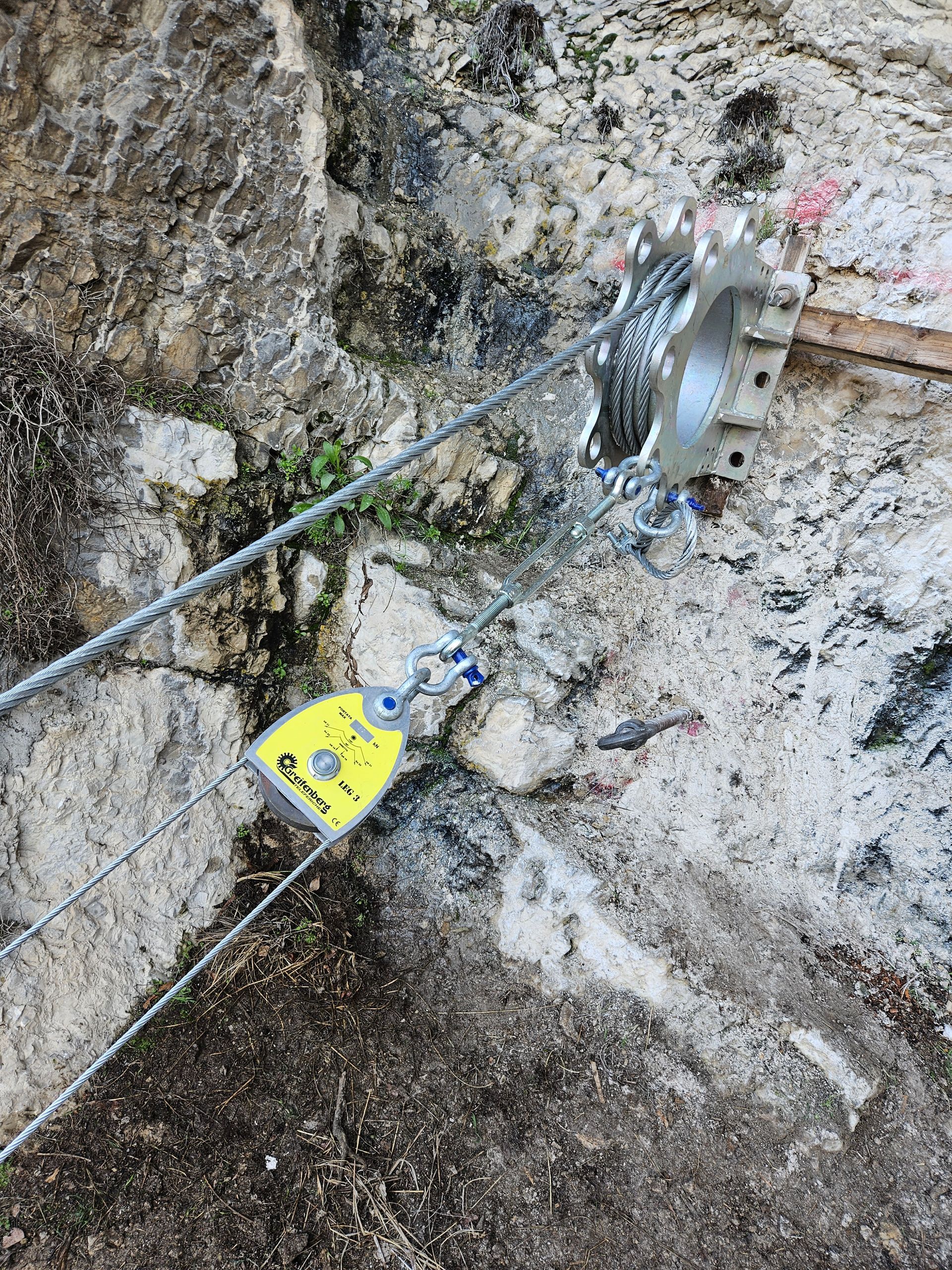 Sistema di carrucole metalliche su una parete rocciosa, comprendente una carrucola, un cavo e un ancoraggio, probabilmente per una teleferica o un'attività simile.
