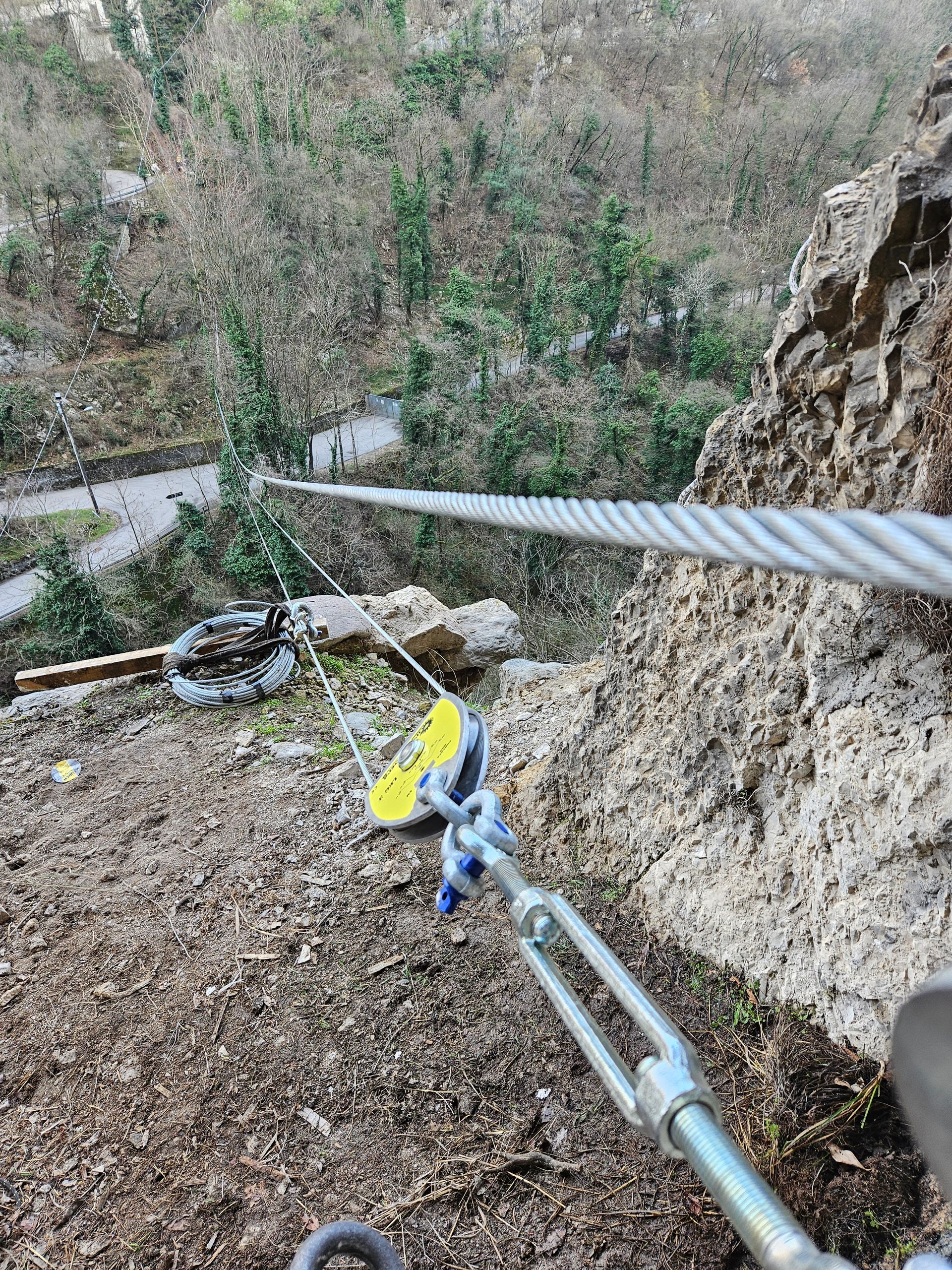 Sistema di cavi ancorato a una scogliera rocciosa, con un filo che si estende verso una lontana linea di alberi e un sentiero.