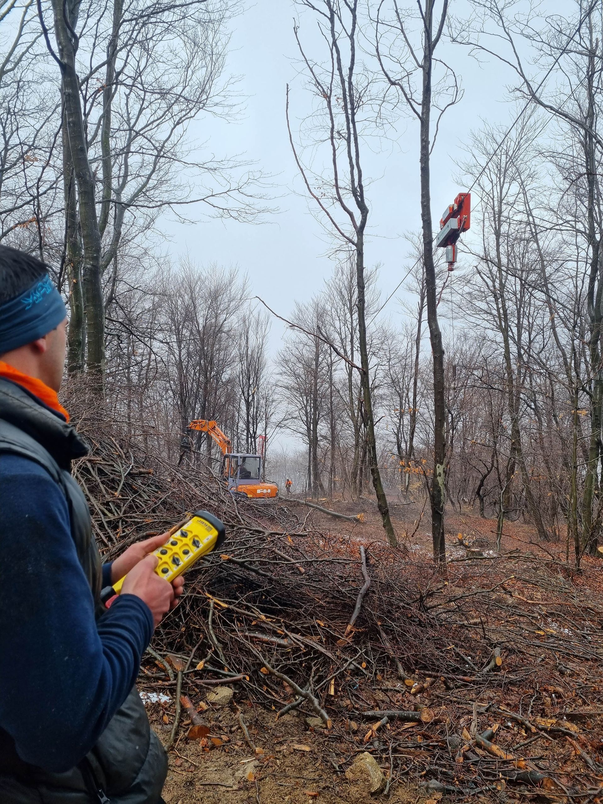 Uomo che aziona un telecomando; l'attrezzatura sta tagliando un albero in una foresta, con un escavatore sullo sfondo.