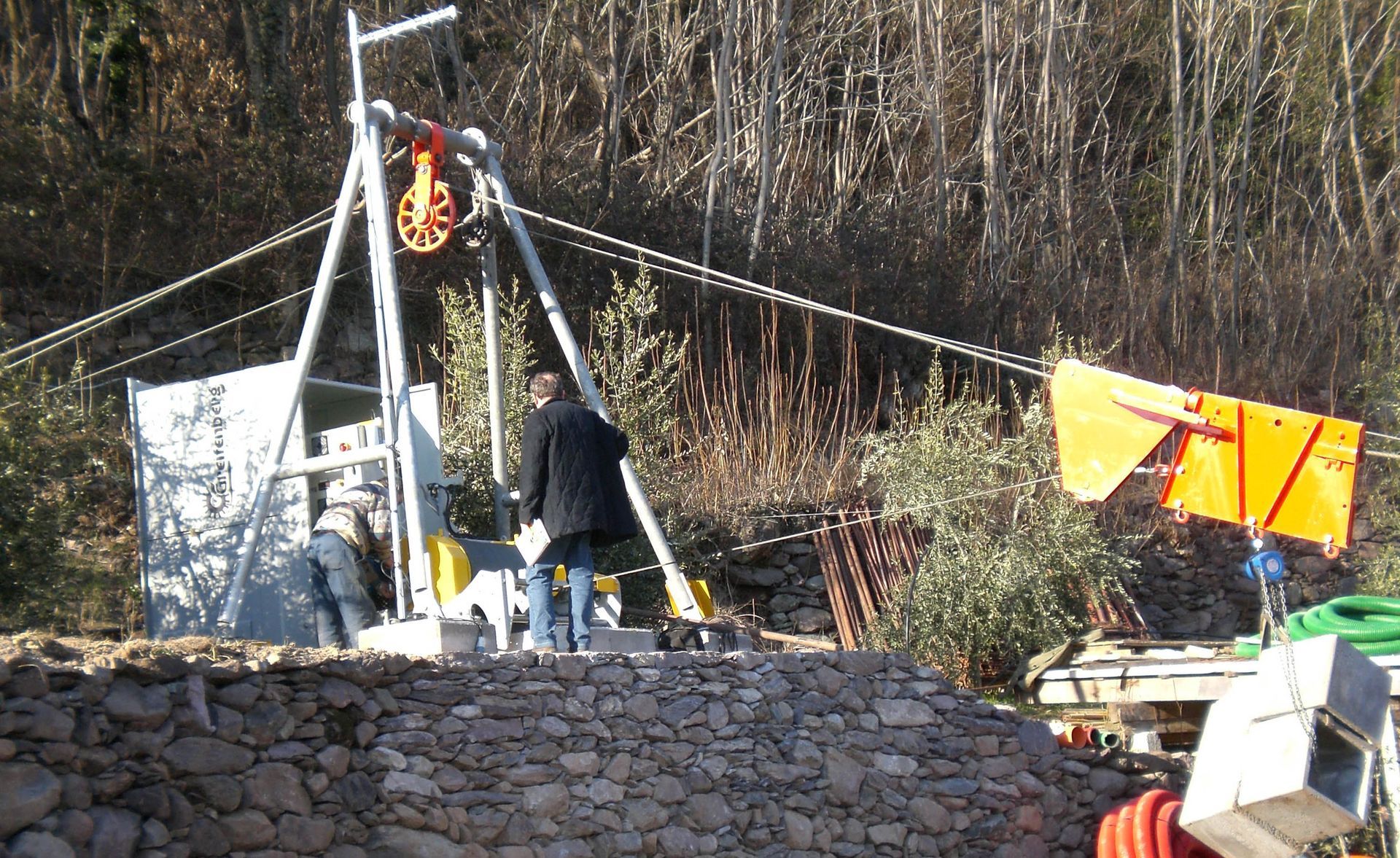 Uomini che azionano un sistema di cavi su un pendio roccioso. Sono visibili macchinari, cavi e alberi arancioni e argentati.