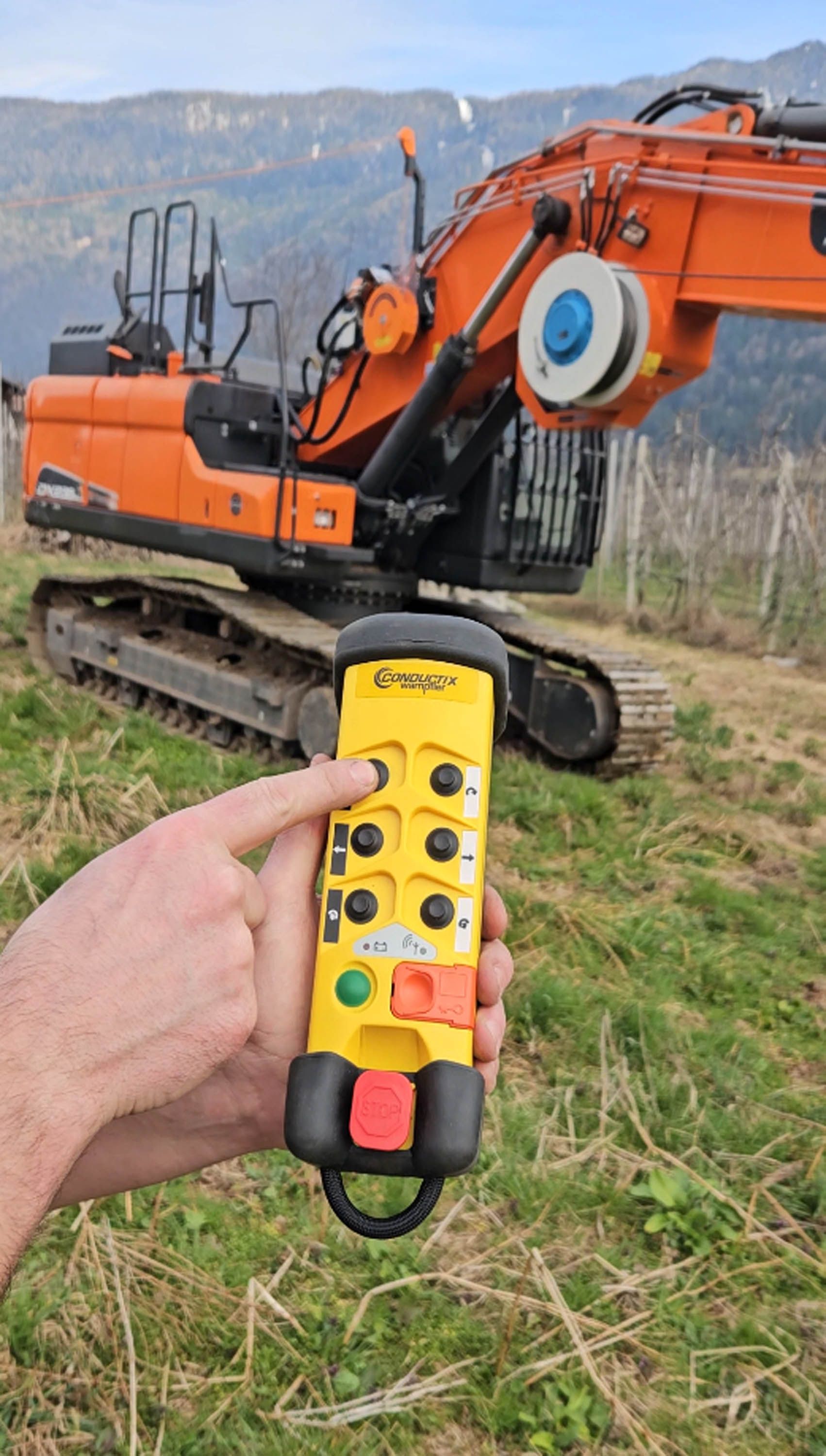 Mano che tiene un telecomando giallo, puntata su un pulsante con un escavatore arancione sullo sfondo e montagne.