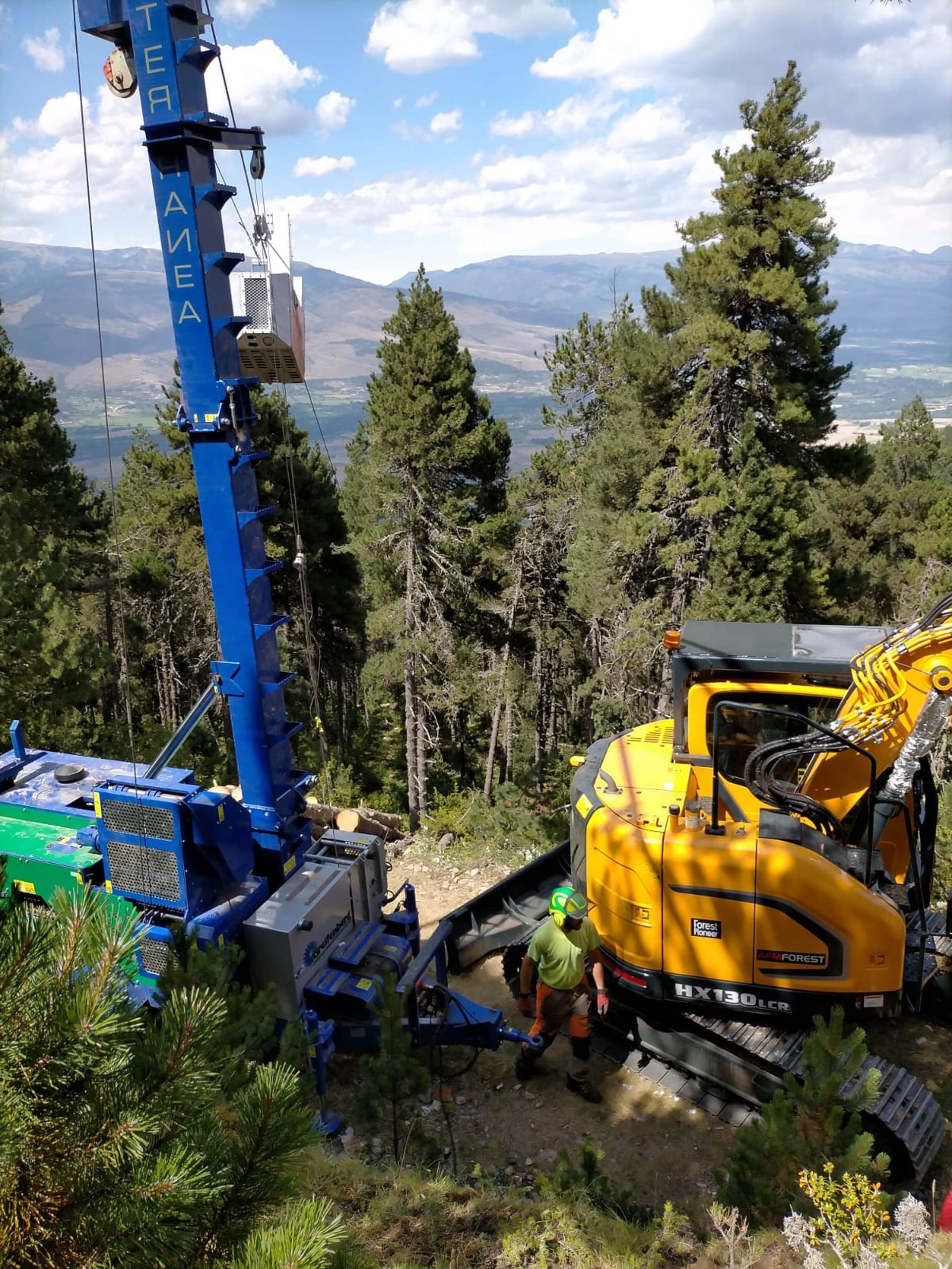 Escavatore giallo e piattaforma aerea blu al lavoro in una zona boschiva; montagne sullo sfondo.