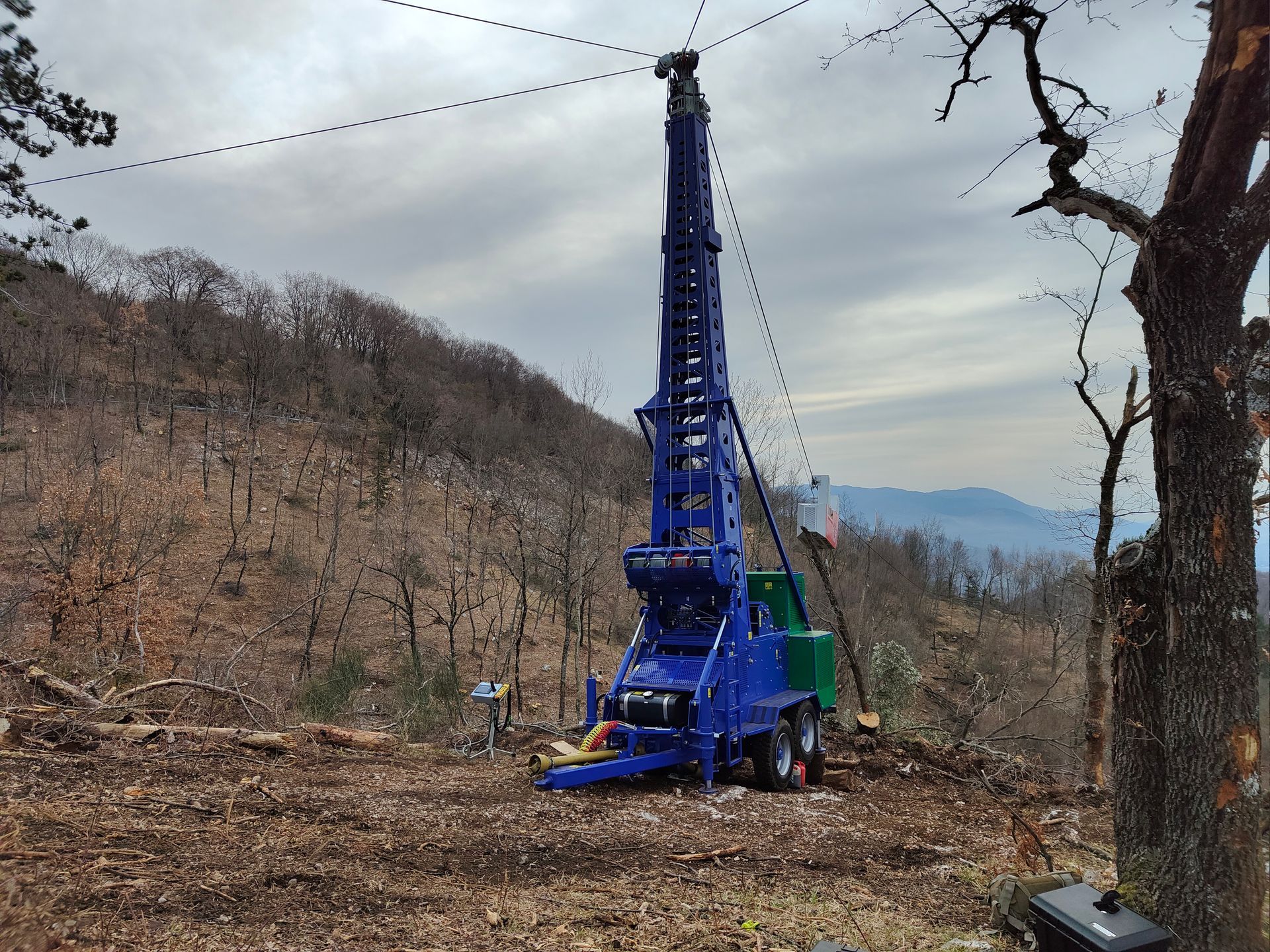 Piattaforma di perforazione blu su una collina, intenta a perforare il terreno. Cavi in alto; alberi e montagne sullo sfondo.
