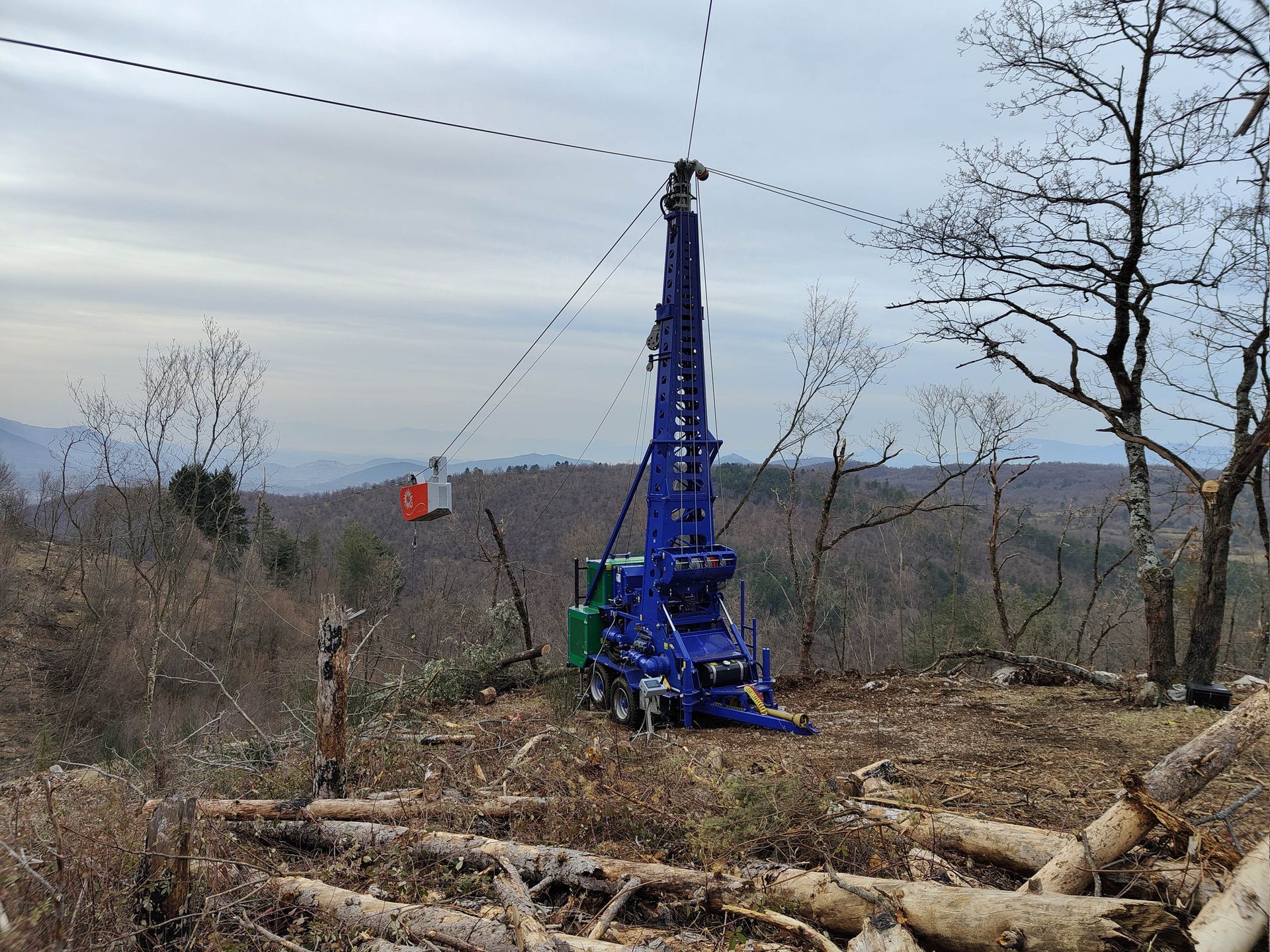 Macchina blu per il taglio del legname con tronchi, cavi in ​​funzione su un'area forestale disboscata. Sullo sfondo montagne nebbiose.