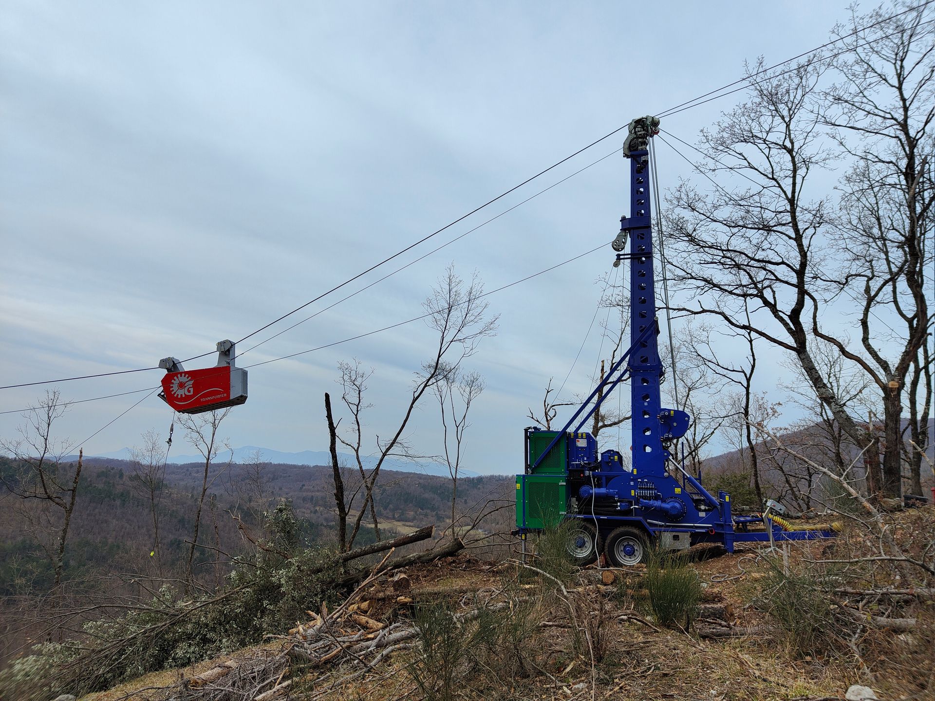 Piattaforma di perforazione blu e carrello rosso su cavi in una zona montuosa e boscosa.