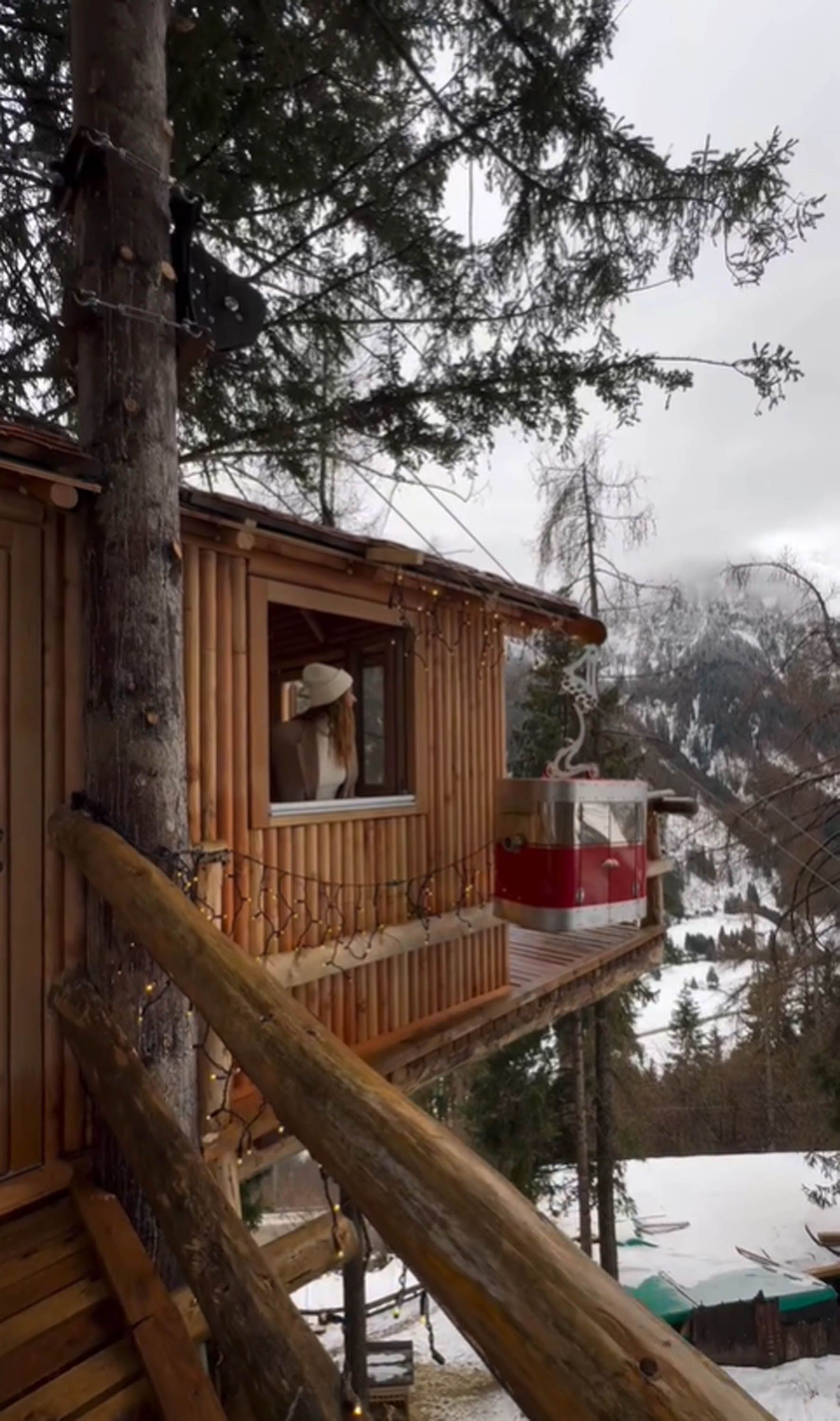 Casa sull'albero in legno con montagne innevate sullo sfondo. Una persona guarda fuori dalla finestra.