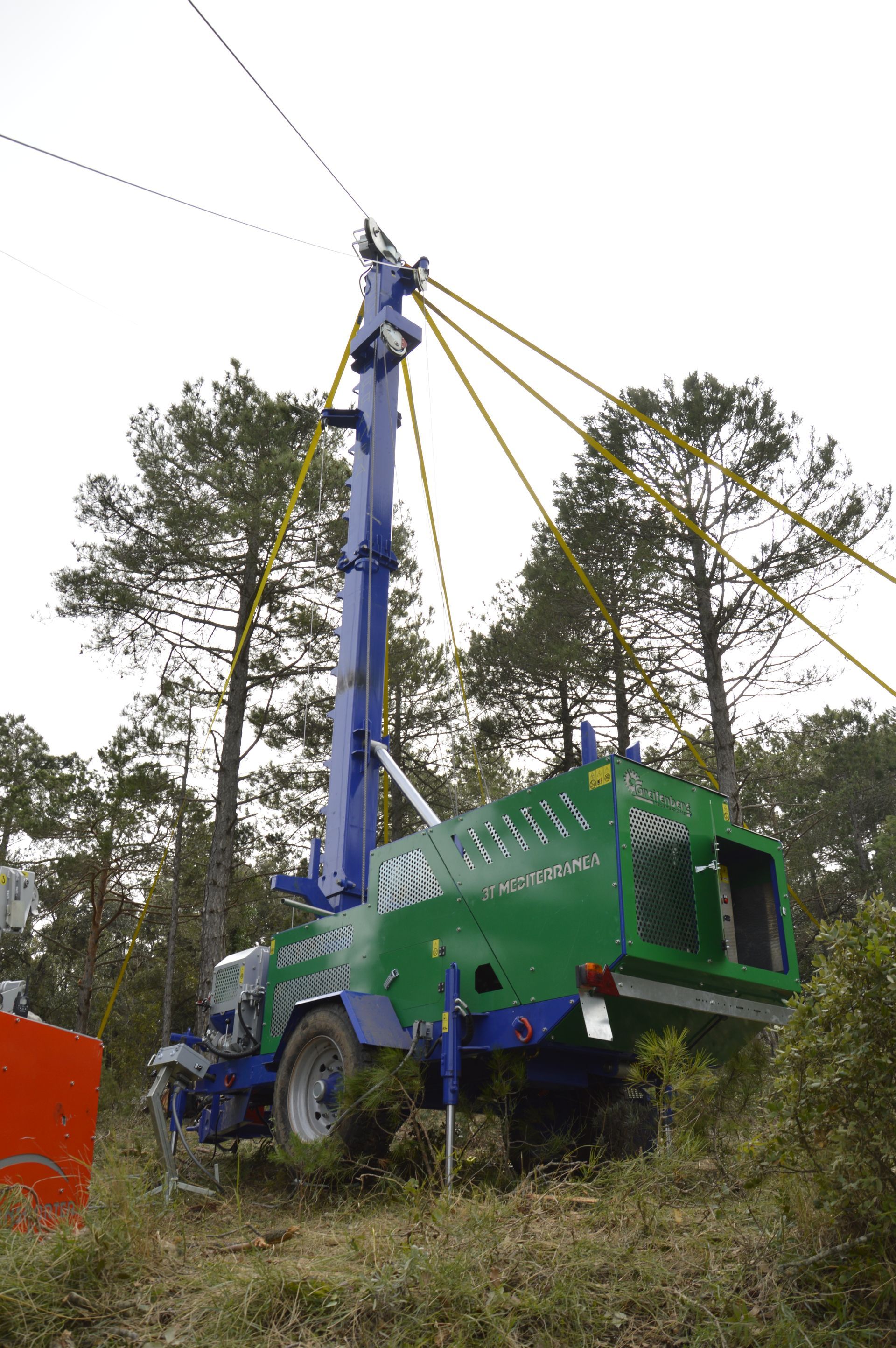 Camion verde e blu con braccio di sollevamento alto in una zona boscosa, a sostegno di linee elettriche.