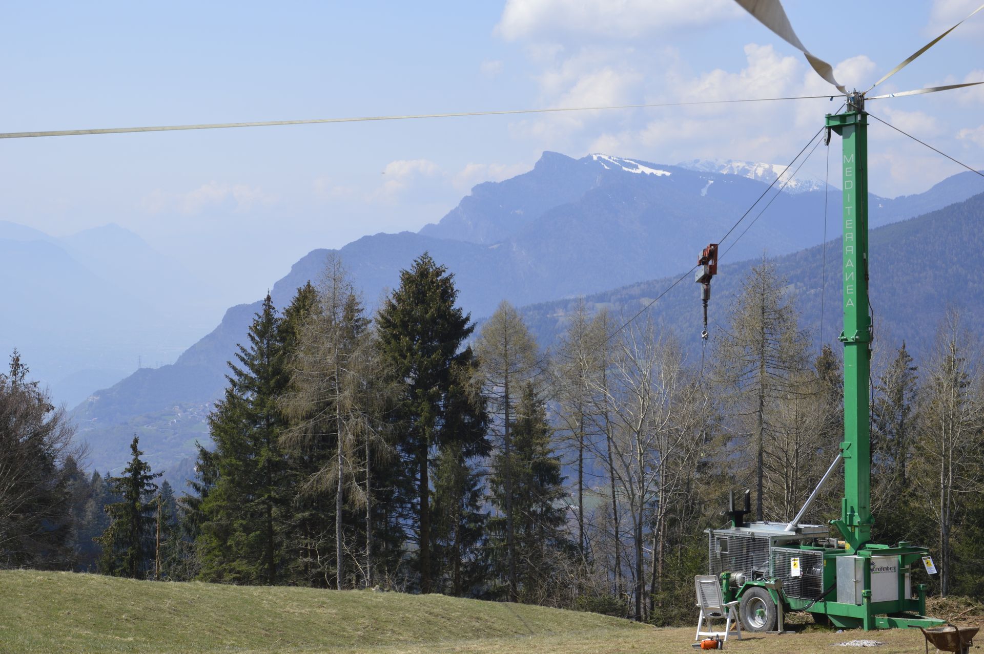 Turbina eolica con vele celesti su un trattore verde su una collina, con montagne e alberi sullo sfondo.