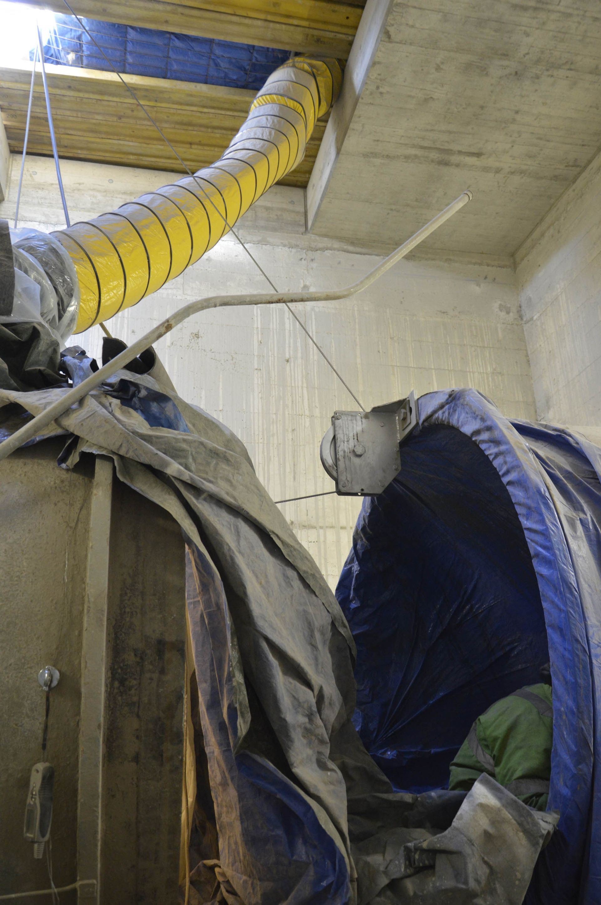 Un cantiere con un condotto di ventilazione giallo, teloni e muri di cemento.