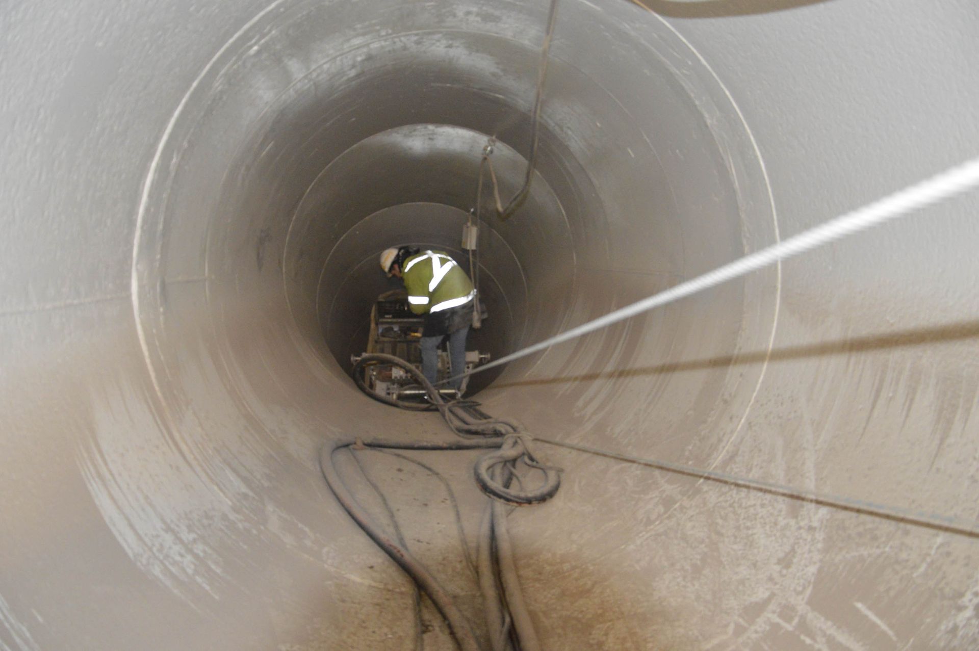 Operaio all'interno di un grande tubo metallico, che pulisce con l'attrezzatura, indossando un giubbotto di sicurezza.