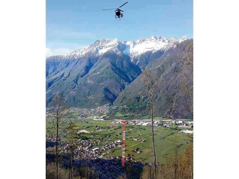 Elicottero che sorvola una valle con una città, una torre rossa e bianca e montagne innevate sullo sfondo.