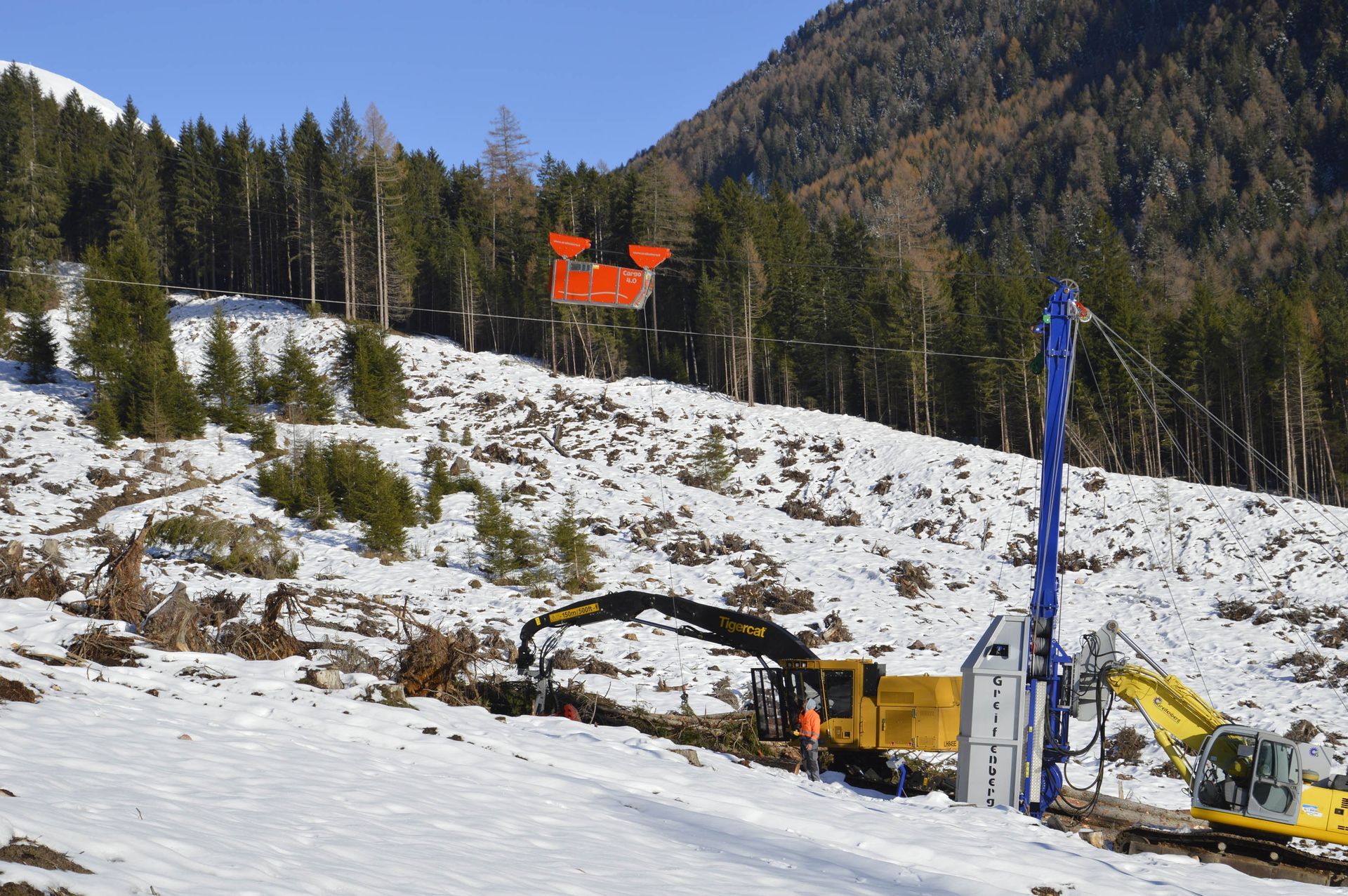 Operazione di disboscamento in montagna innevata; macchinari pesanti, container arancione sospeso sopra.