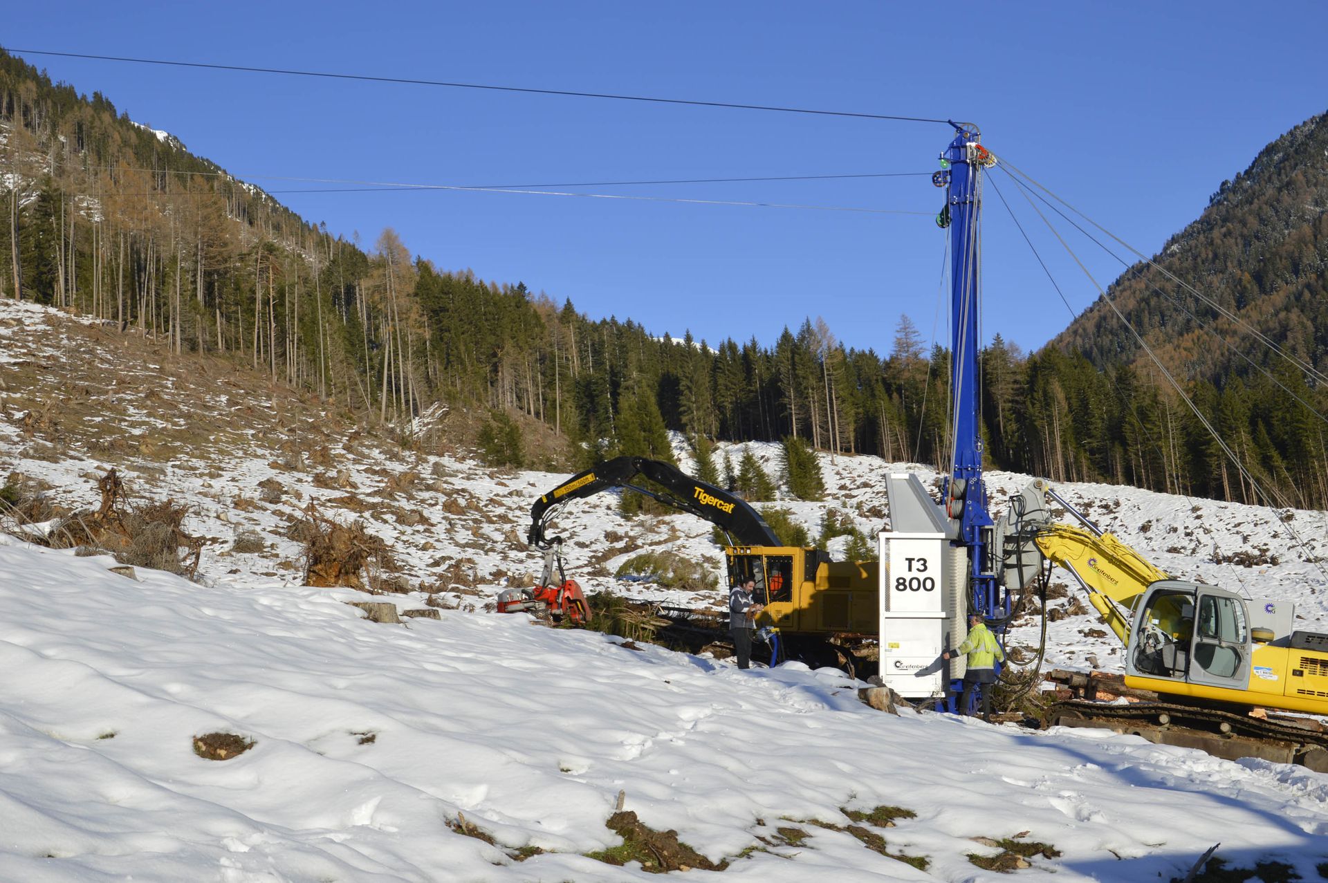 Cantiere in una foresta innevata: escavatore, gru e macchinari gialli al lavoro su un pendio sotto i cavi elettrici.