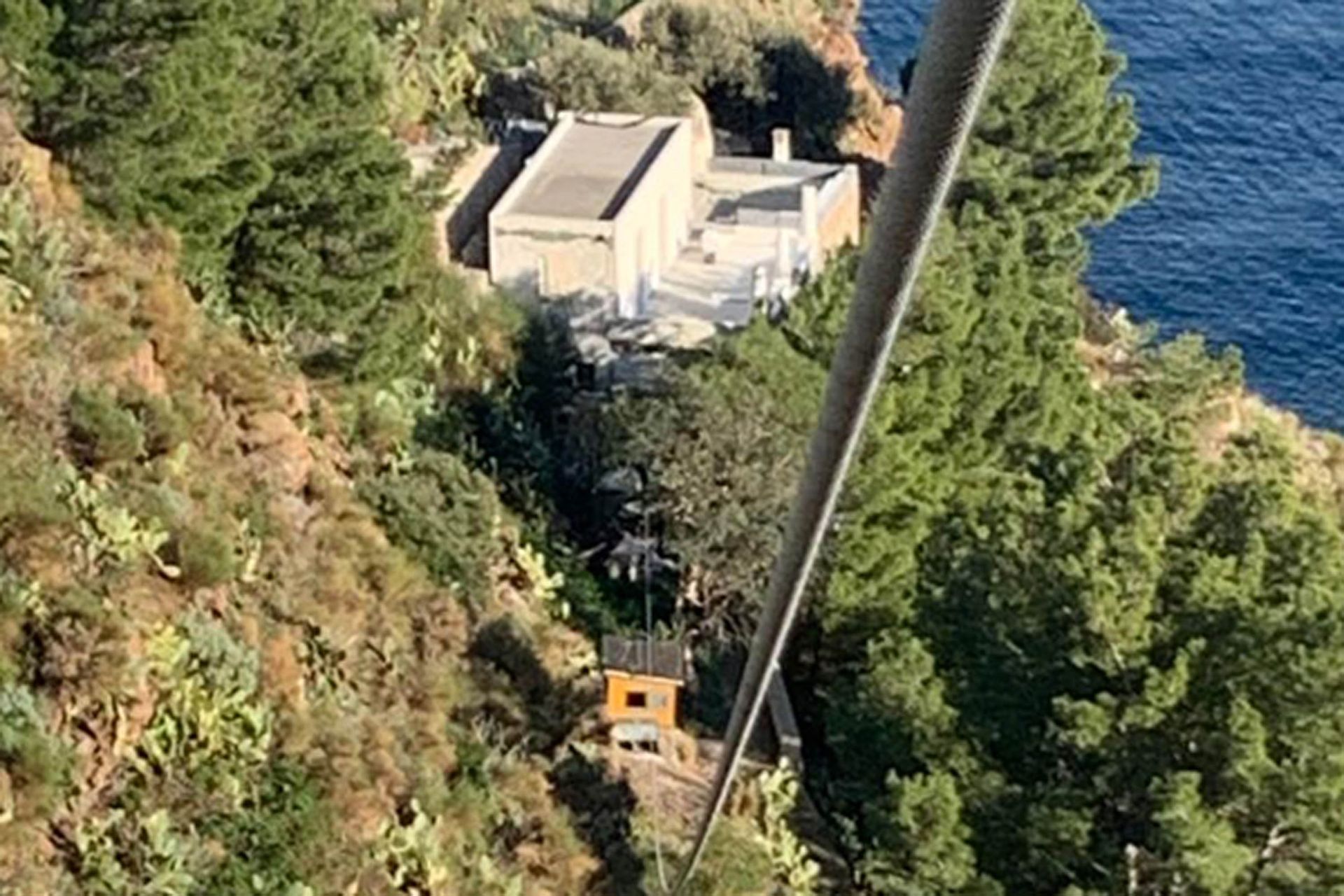 Vista aerea di una funivia che attraversa una scogliera, con un edificio e il mare visibili.