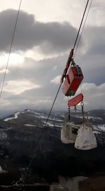 Funivia rossa e bianca con secchi che trasportano materiali, sopra una catena montuosa innevata sotto un cielo nuvoloso.