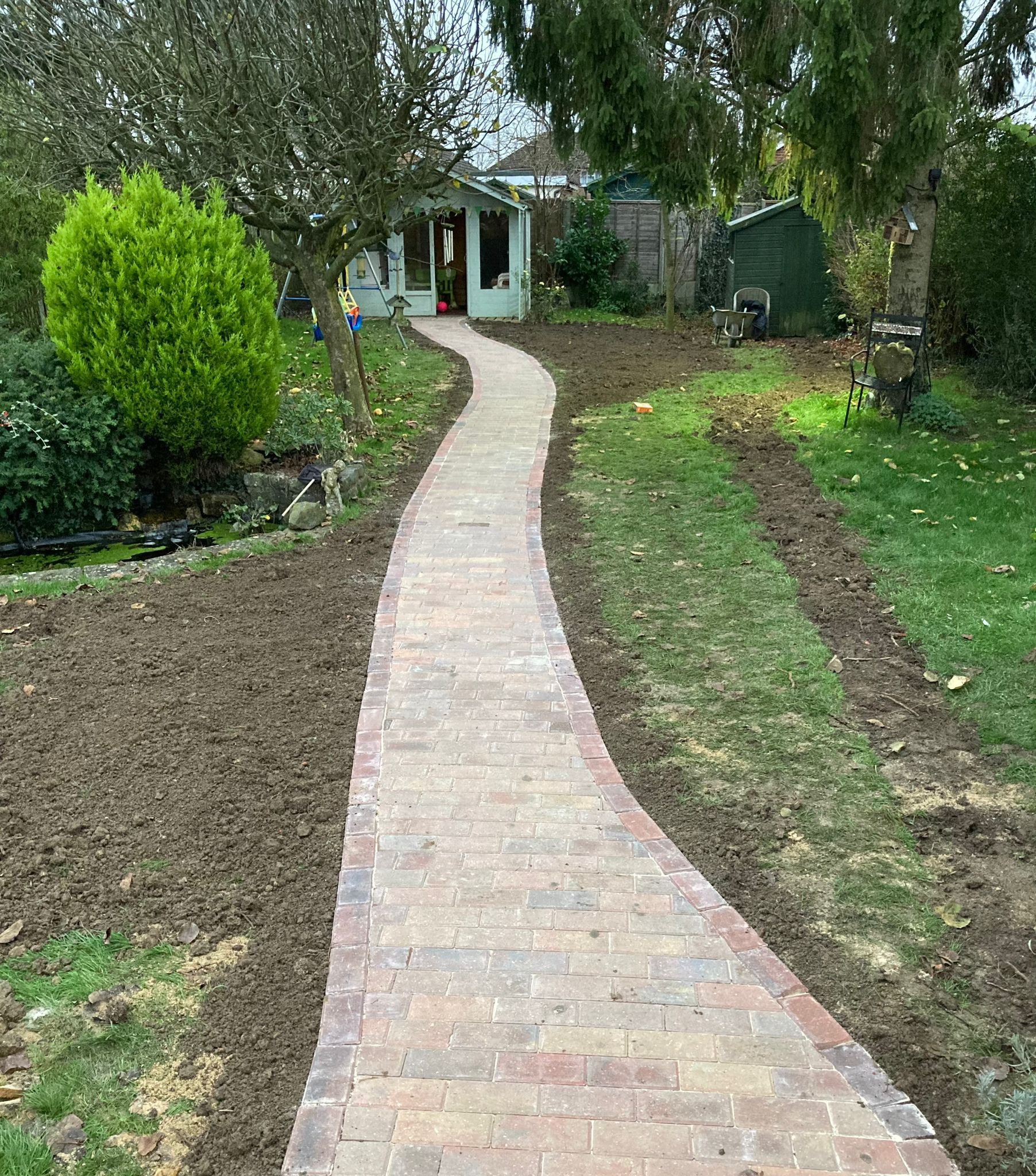 Block paved path through a garden with partly laid lawn and a shed