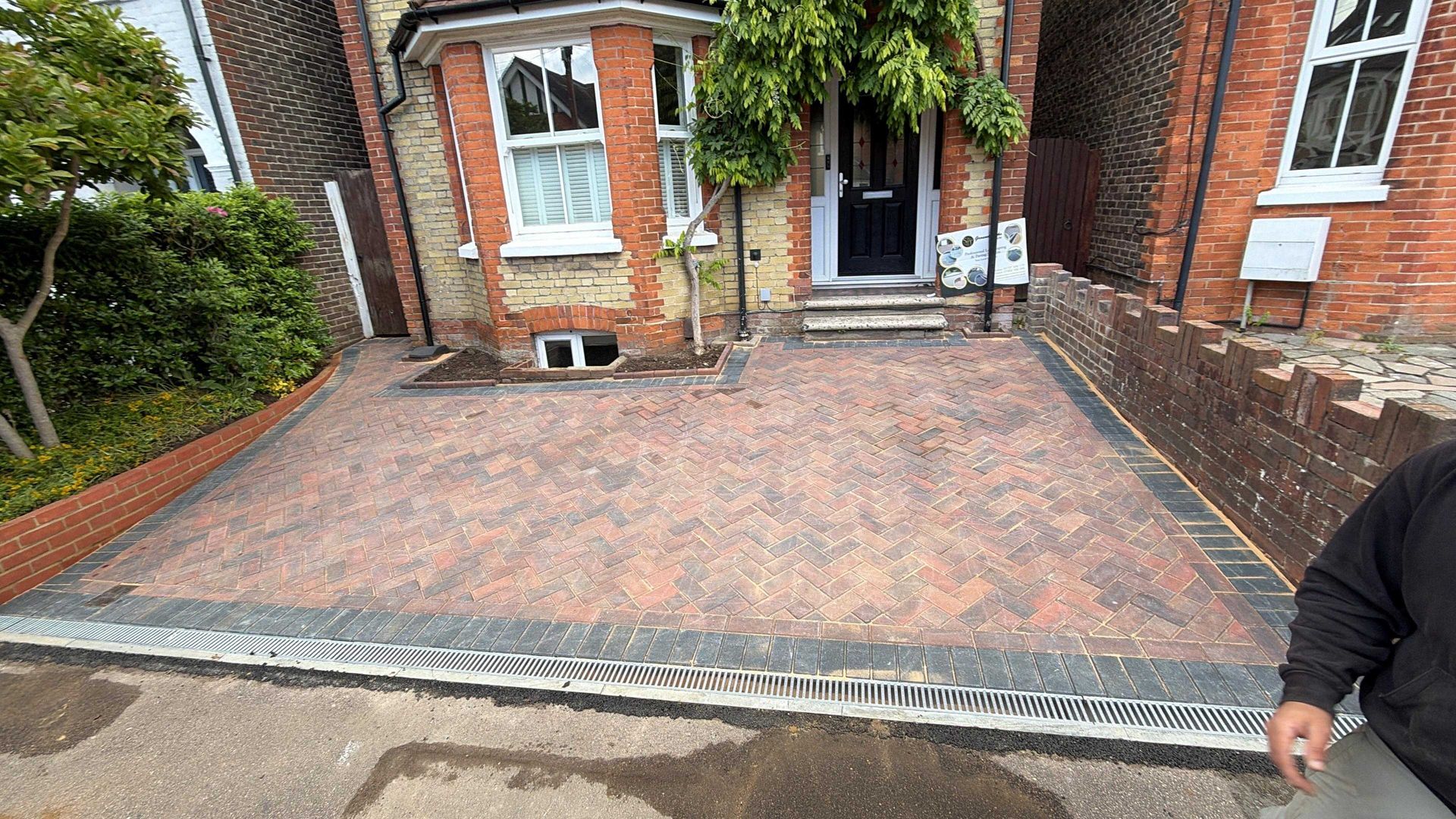 Brick driveway laid with red pavers in front of a brick house, bordered by a curb and garden greenery.