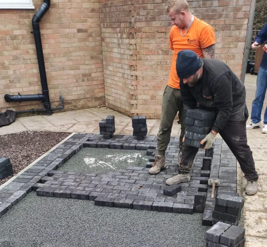Two men laying dark paving stones in a backyard, beside a brick wall, with another person partially visible.