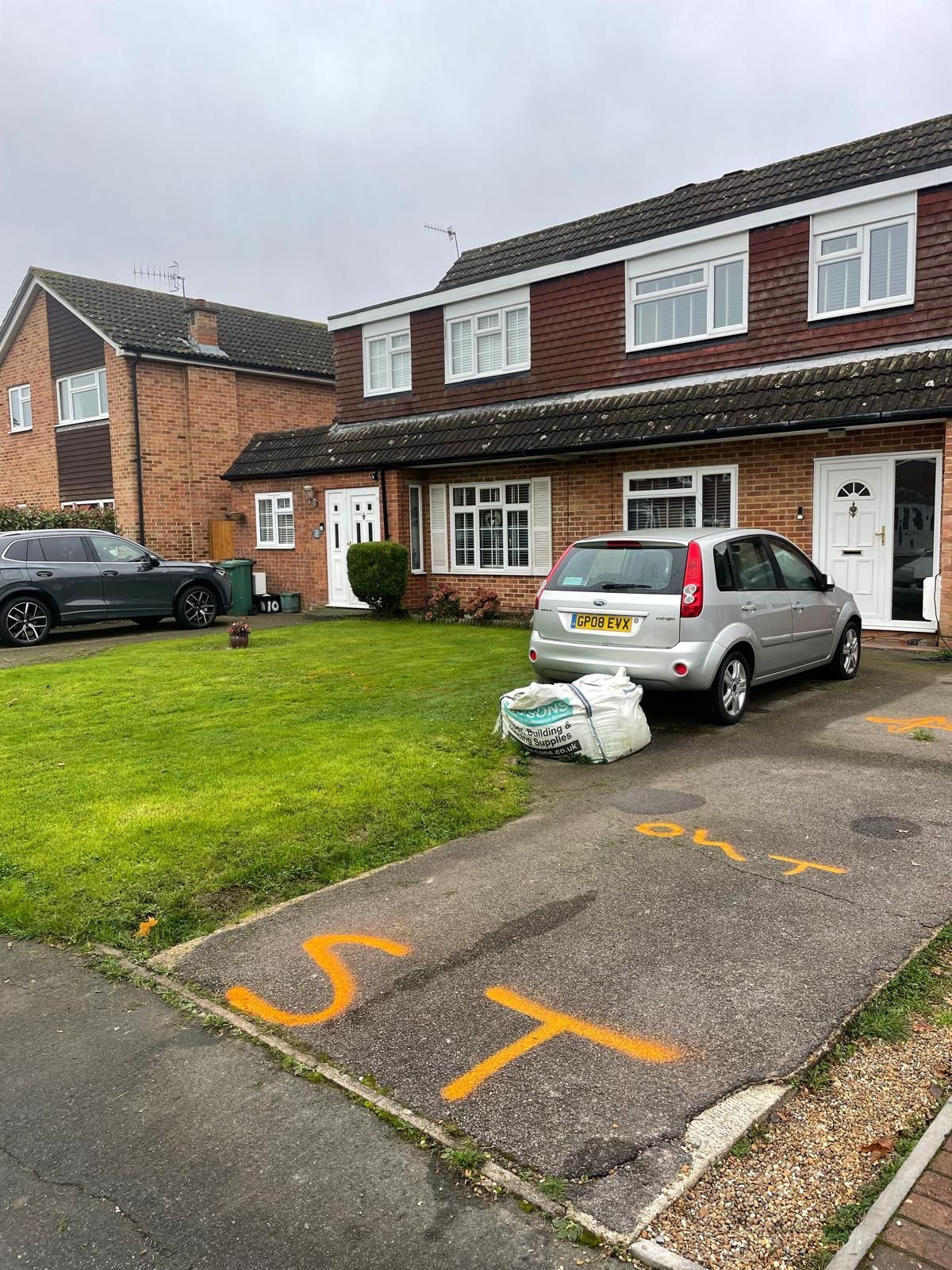 Row houses with cars parked in driveways, yard with trash bags, orange markings on pavement.