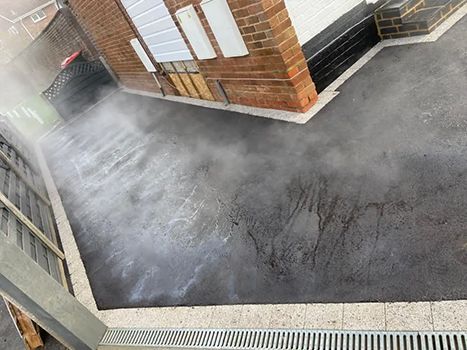 A black asphalt driveway with a drain and a brick building in the background.